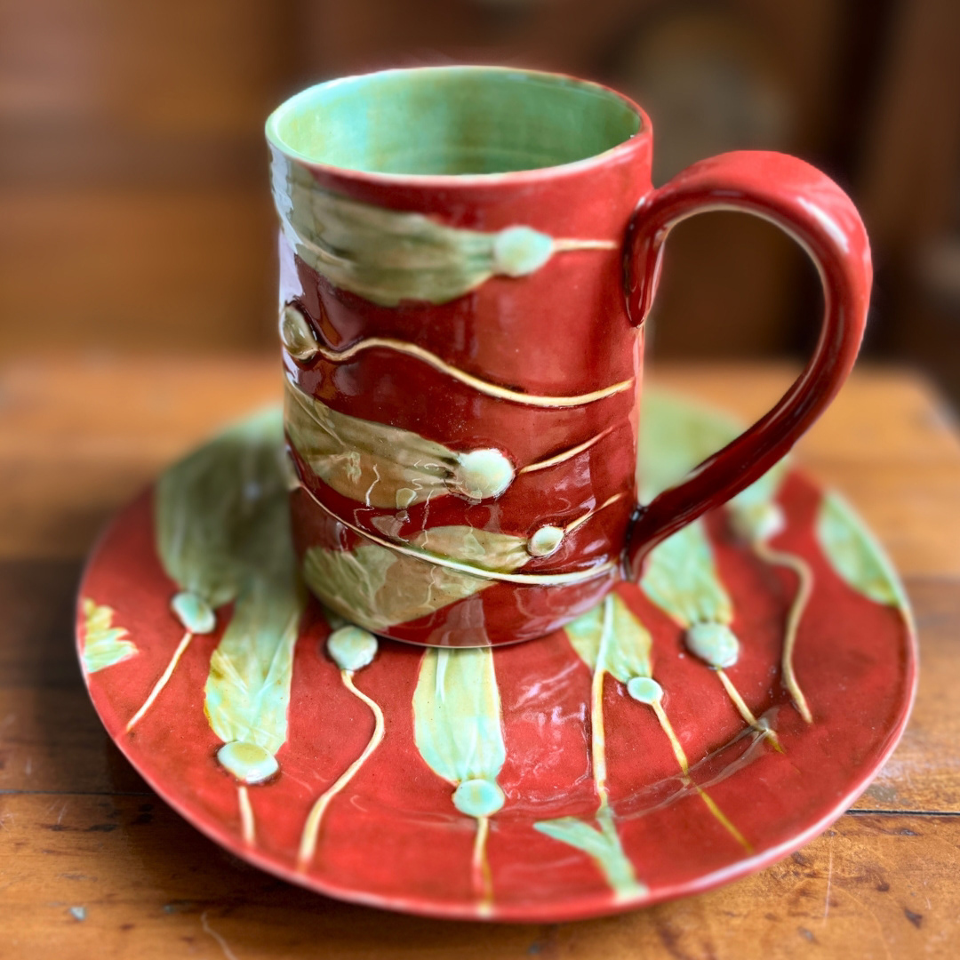 A stein-style mug resting on a matching plate, both adorned with imprints of green bullwhip kelp, draping across the surfaces. in shades of red and greens