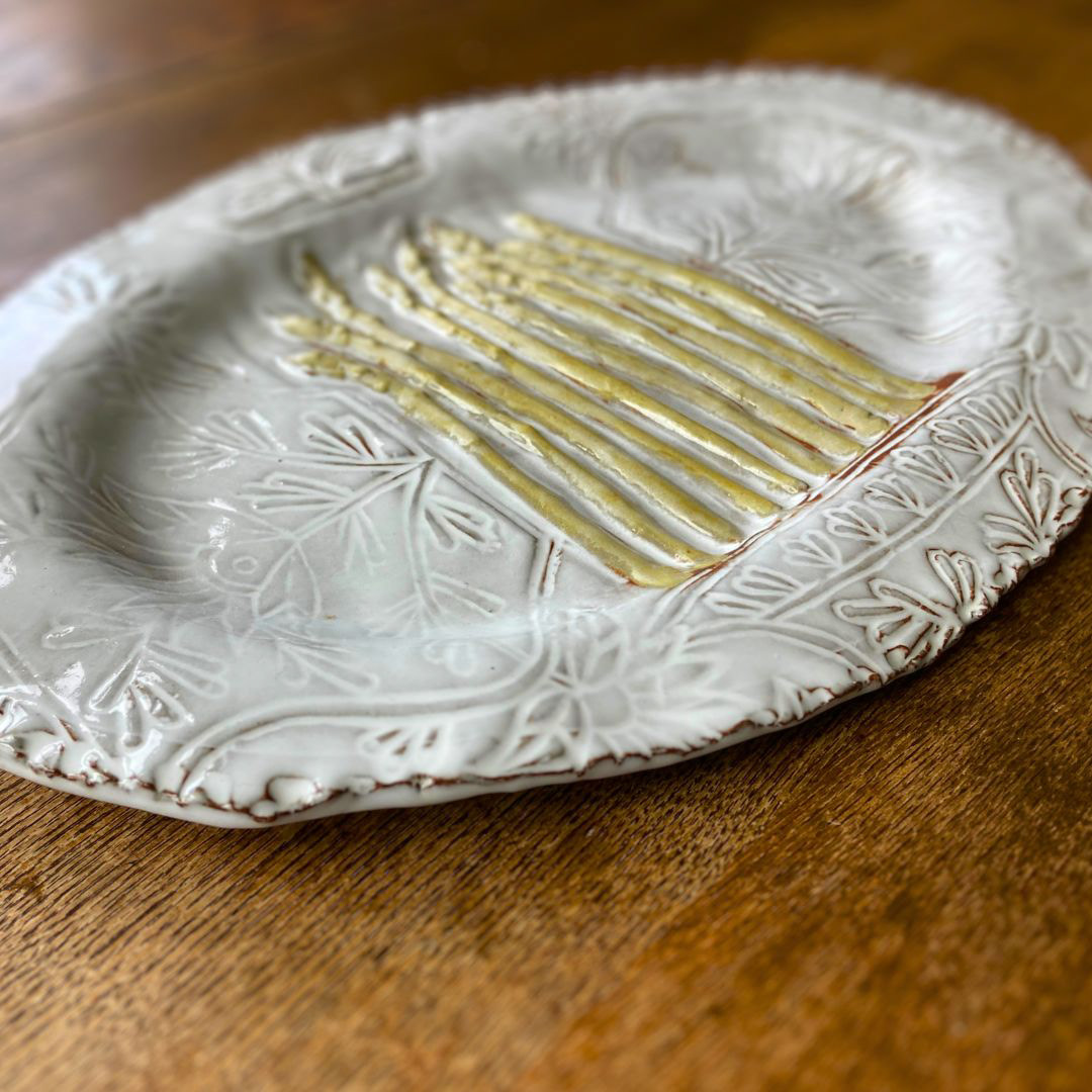 Full view of a handmade ceramic platter on a wooden table, featuring raised asparagus details over a delicate white floral pattern.