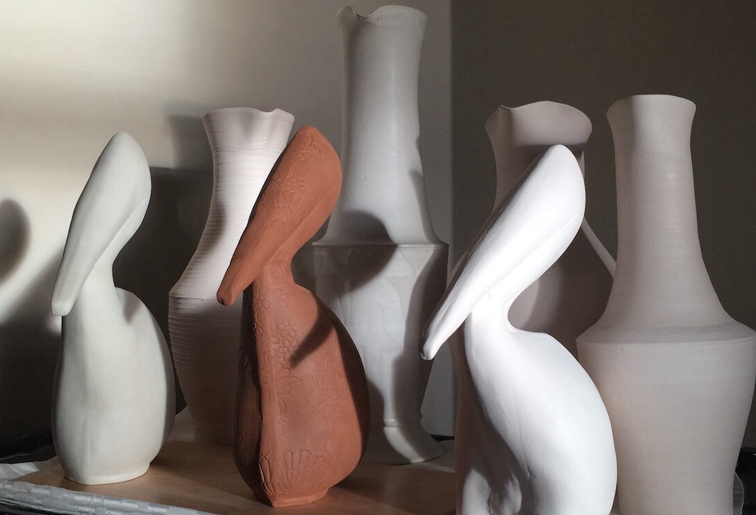 three pelican sculptures in shaeds of white and reddish clay color. in the back four tall white handmade vases are visible against wall.