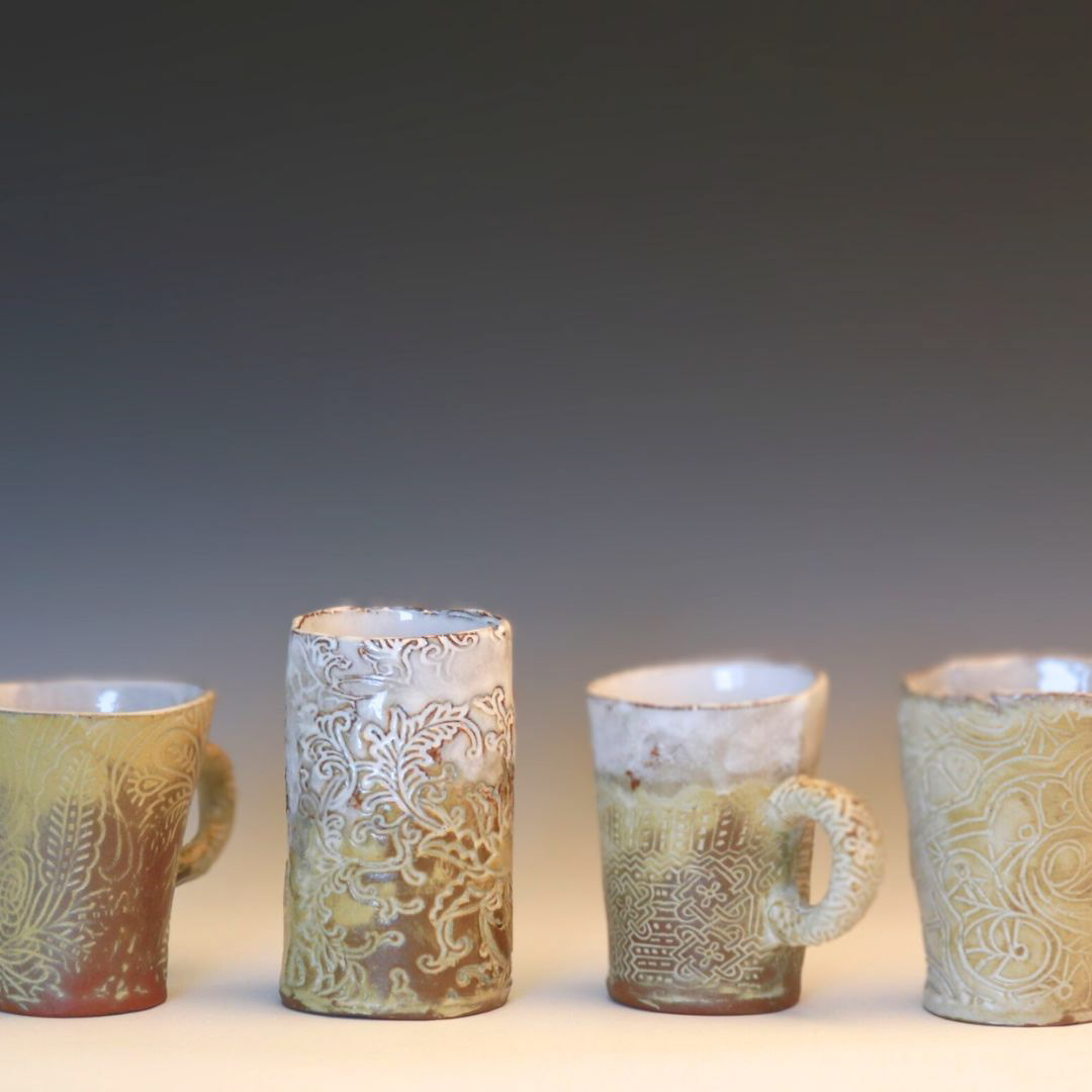 Four handmade ceramic mugs with de-bossed floral patterns in shades of white, tan and gold.