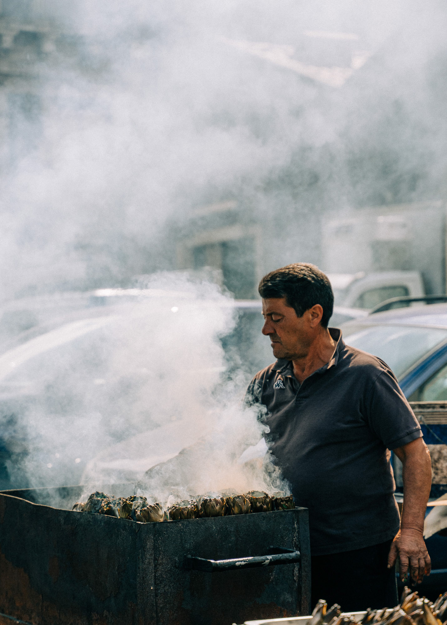 Street photography showing a man grilling artichokes under heavy smoke at the market of Catania