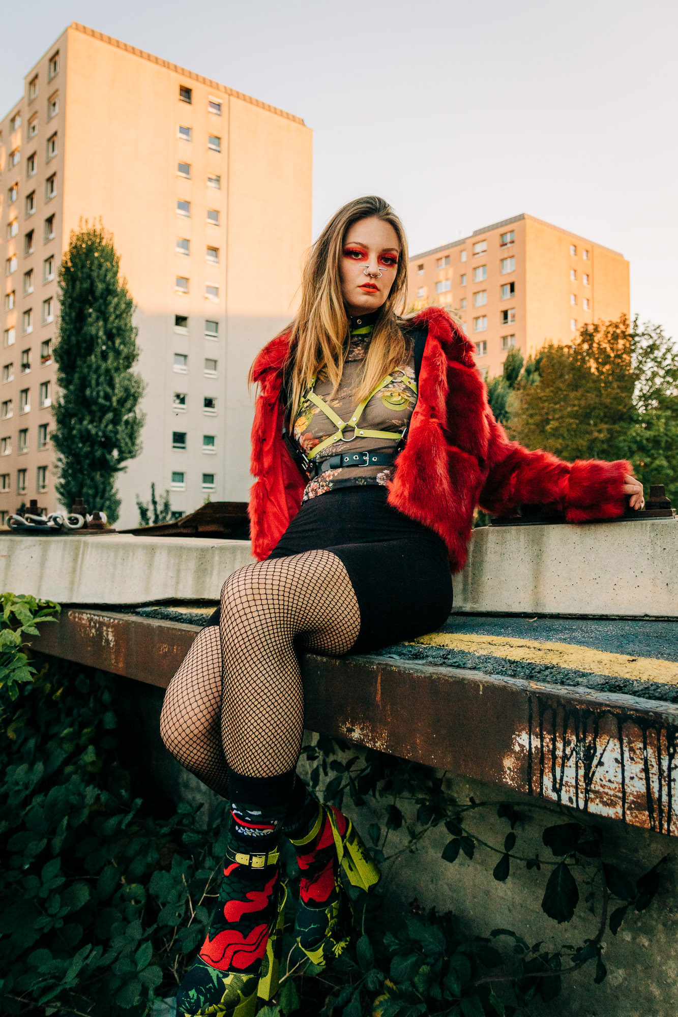 Portrait of a girl with red makeup, a red fur coat, a leather harness and high heels sitting at an industrial area