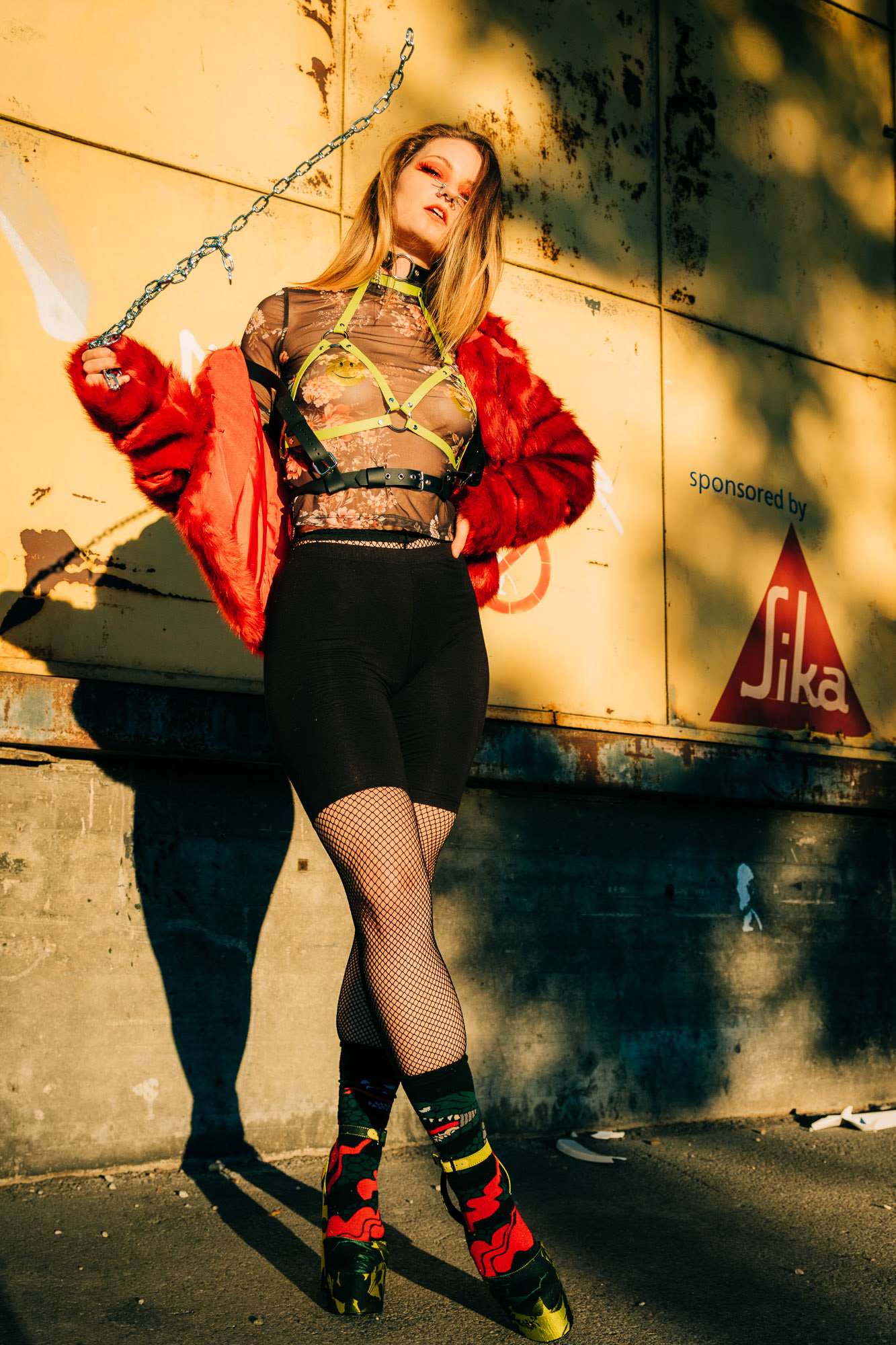 Fashion portrait of a girl wearing high heels, leather harness and a red fur coat while swinging an iron chain in her hand