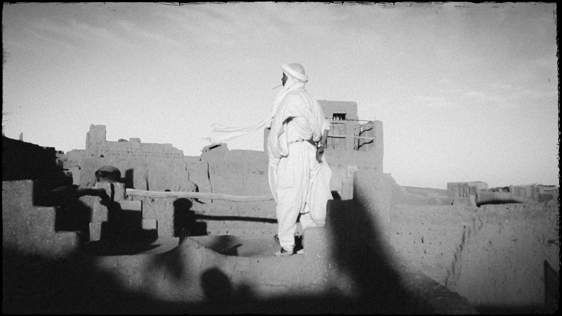 Balck and white portrait of a Berber man standing on top of a clay hut as his turban is waving in the wind
