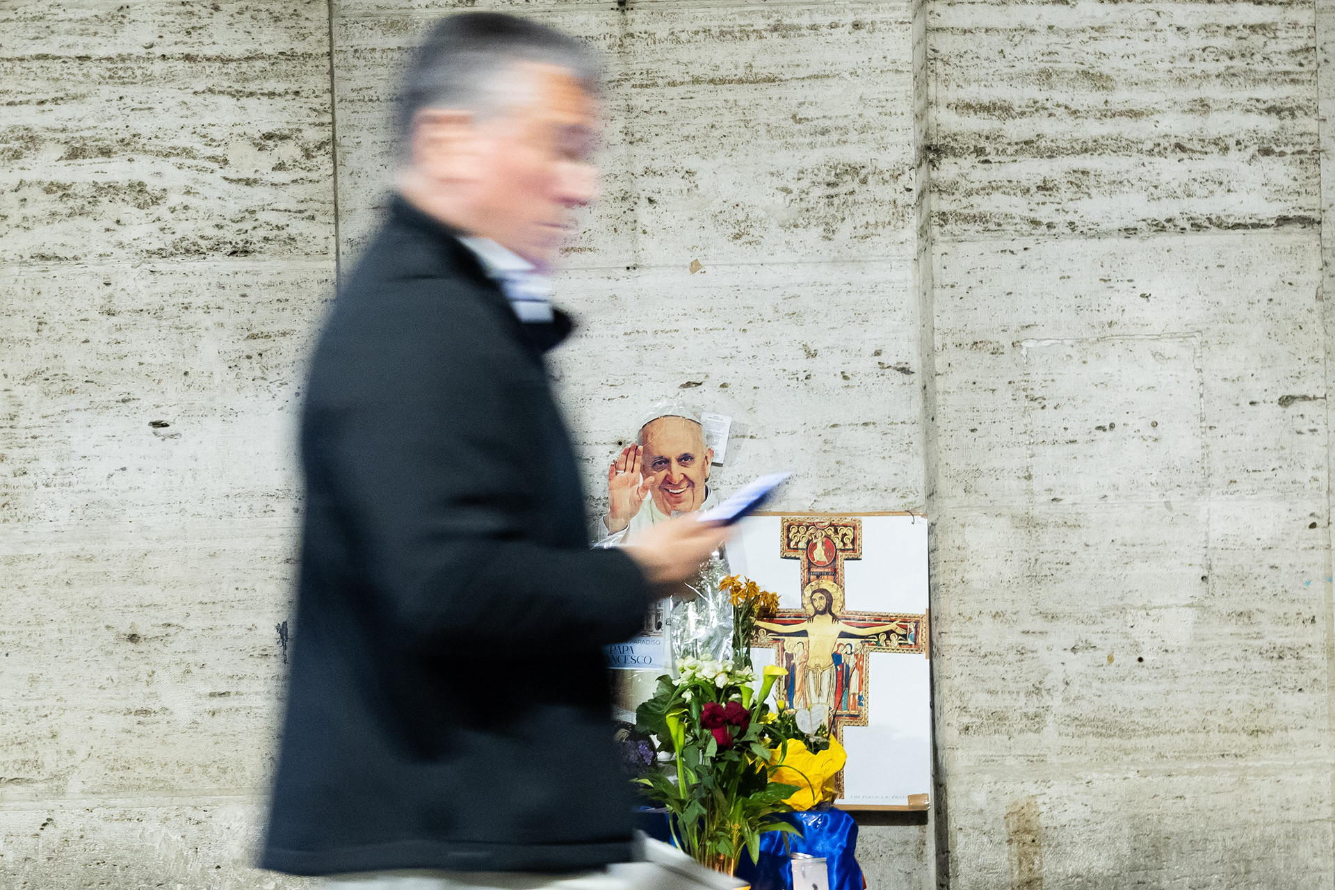 Memorial to Pope Francis near St. Peter’s Square, Rome. A man walks focused on his phone; behind him, a portrait of the smiling Pope. Photo © Marek Ladzinski / FORUM