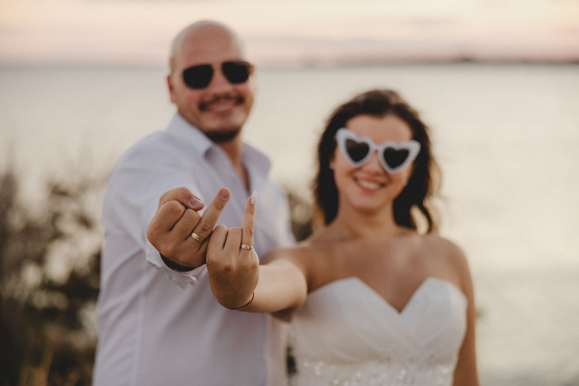 Забавна и непринудена сватбена снимка на младоженци, показващи пръст с брачни халки на морския бряг. Модерен сватбен фотограф Несебър.