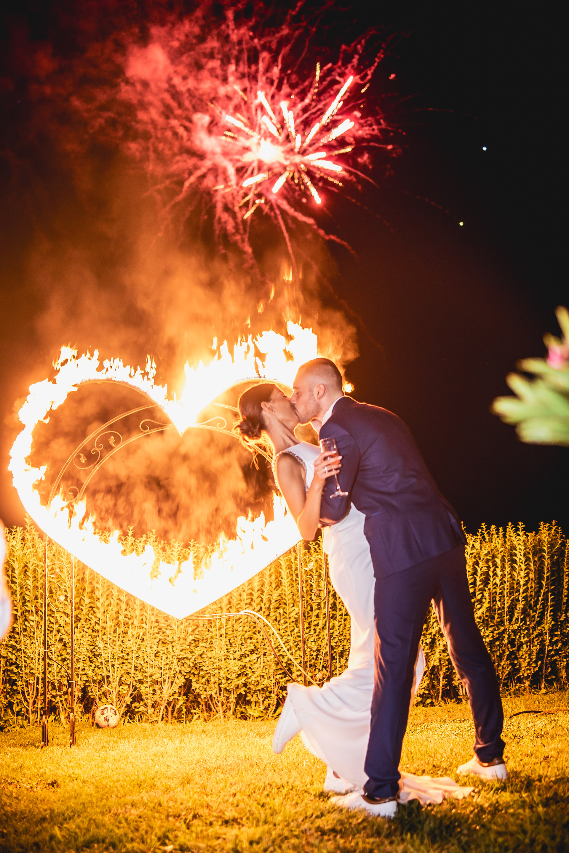 Младоженец целува младоженка, която го е прегърнала и държи чаша в ръка. На заден фон горящо сърце и фойерверки озаряват нощта, създавайки романтична и магична атмосфера