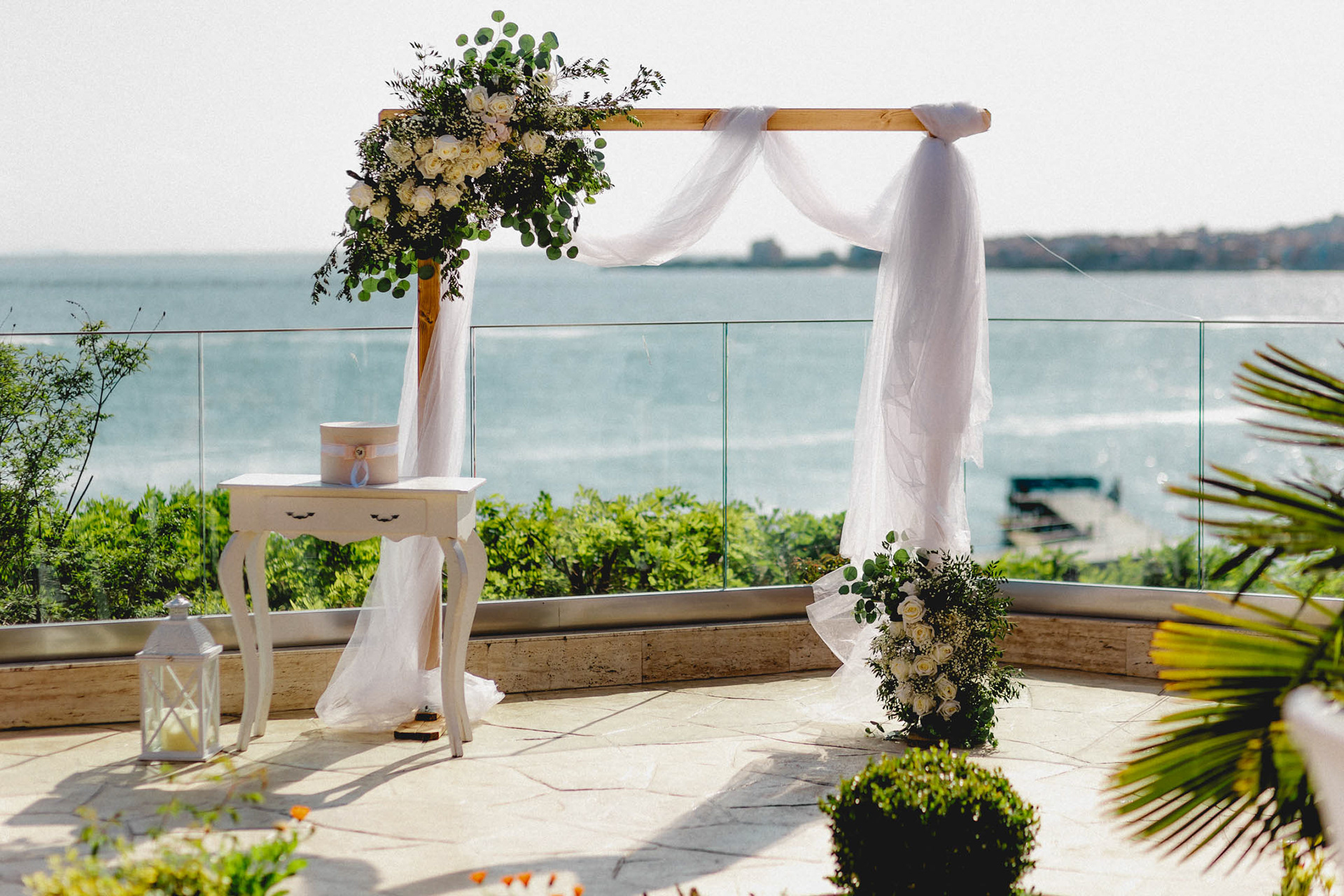 Правоъгълна дървена сватбена арка с бели цветя и тюл на тераса с панорамен изглед към морето в Равда