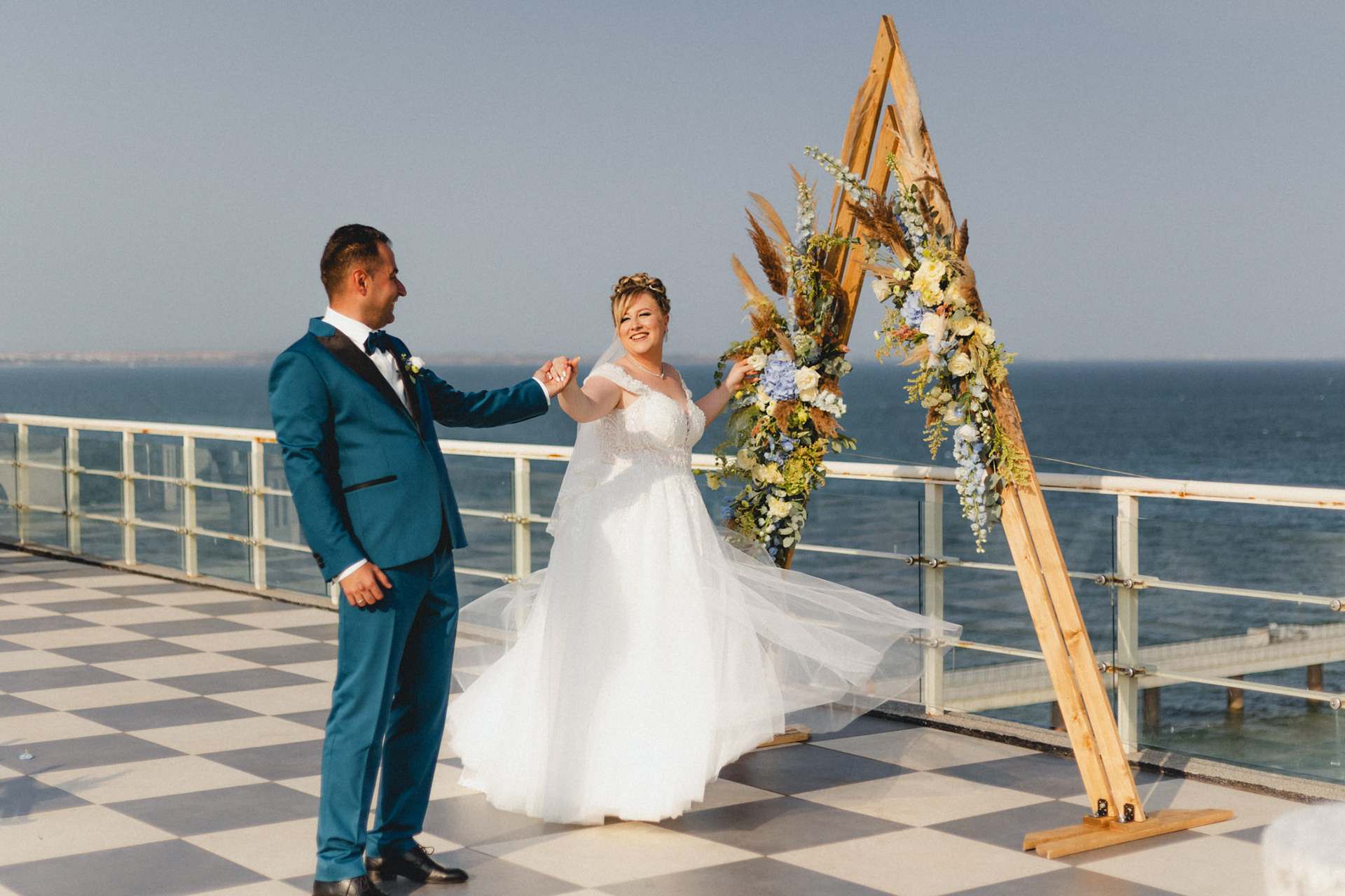 Младоженци танцуват пред дървена сватбена арка с бохо украса на терасата на Морското казино с гледка към морето в Бургас