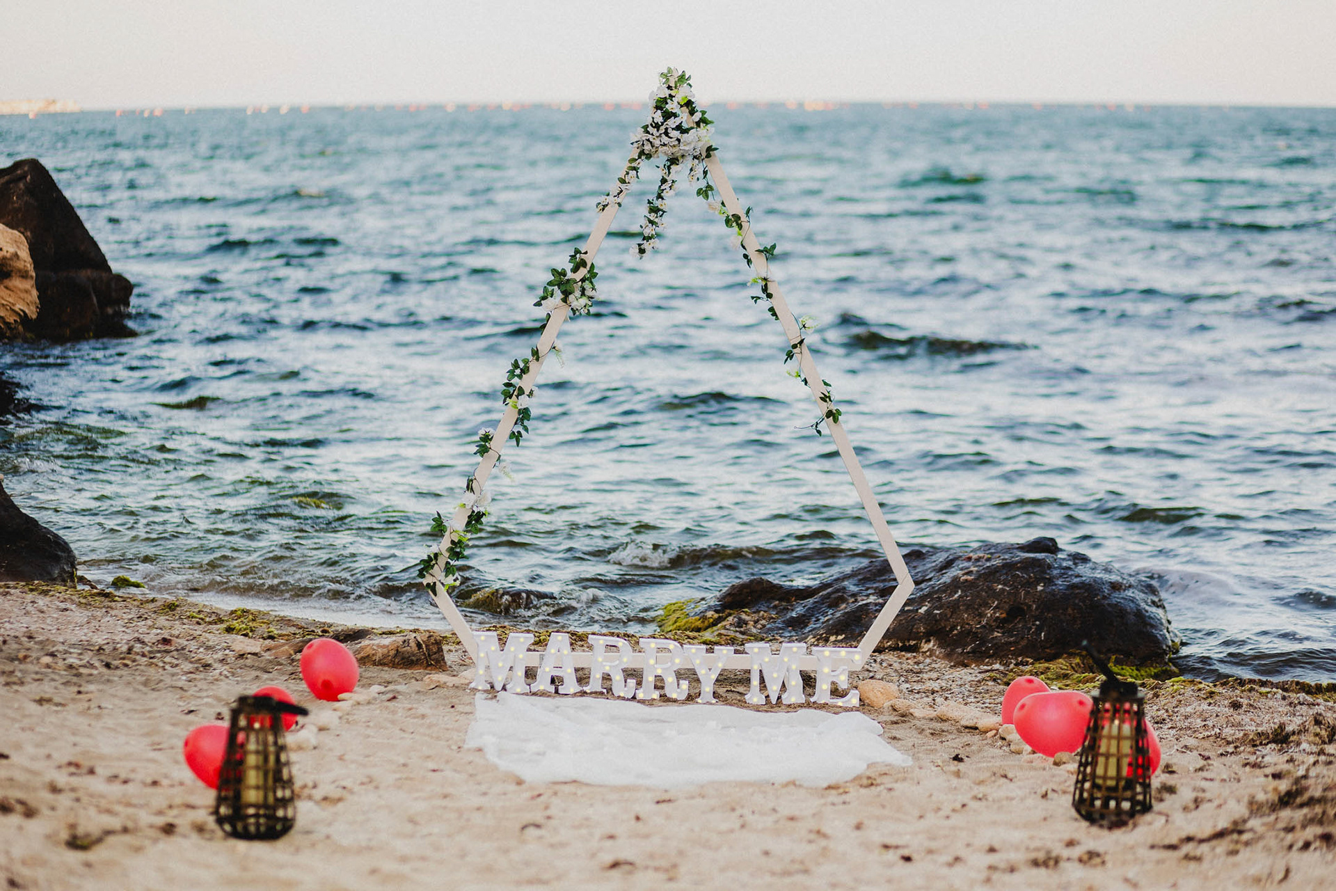 Бяла арка във формата на диамант, украсена с цветя и надпис "Marry Me", поставена на морския бряг в Созопол за романтично предложение за брак