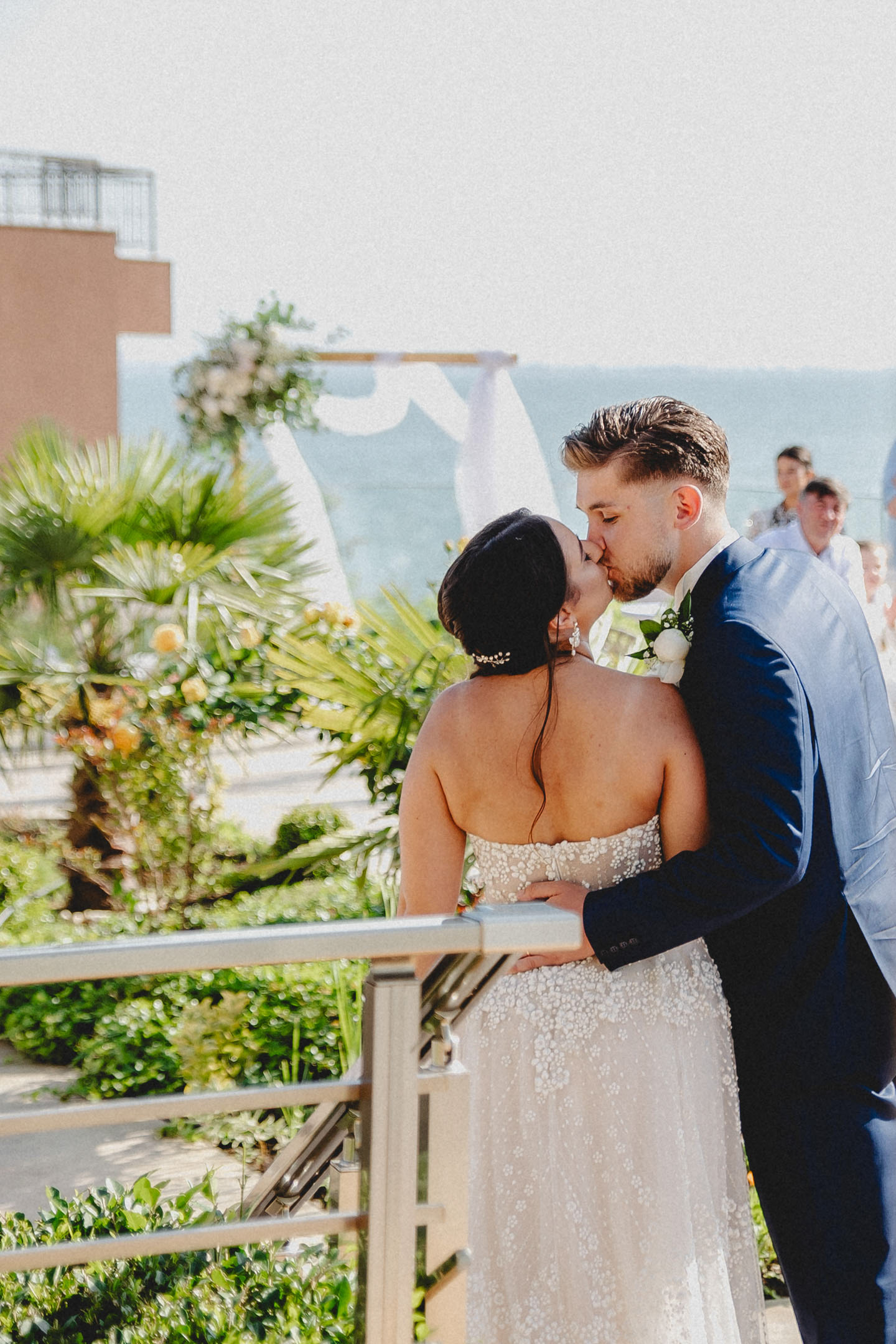 Младоженци се целуват пред сватбената арка с морски изглед към Ахелой, заобиколени от зеленина и гости.