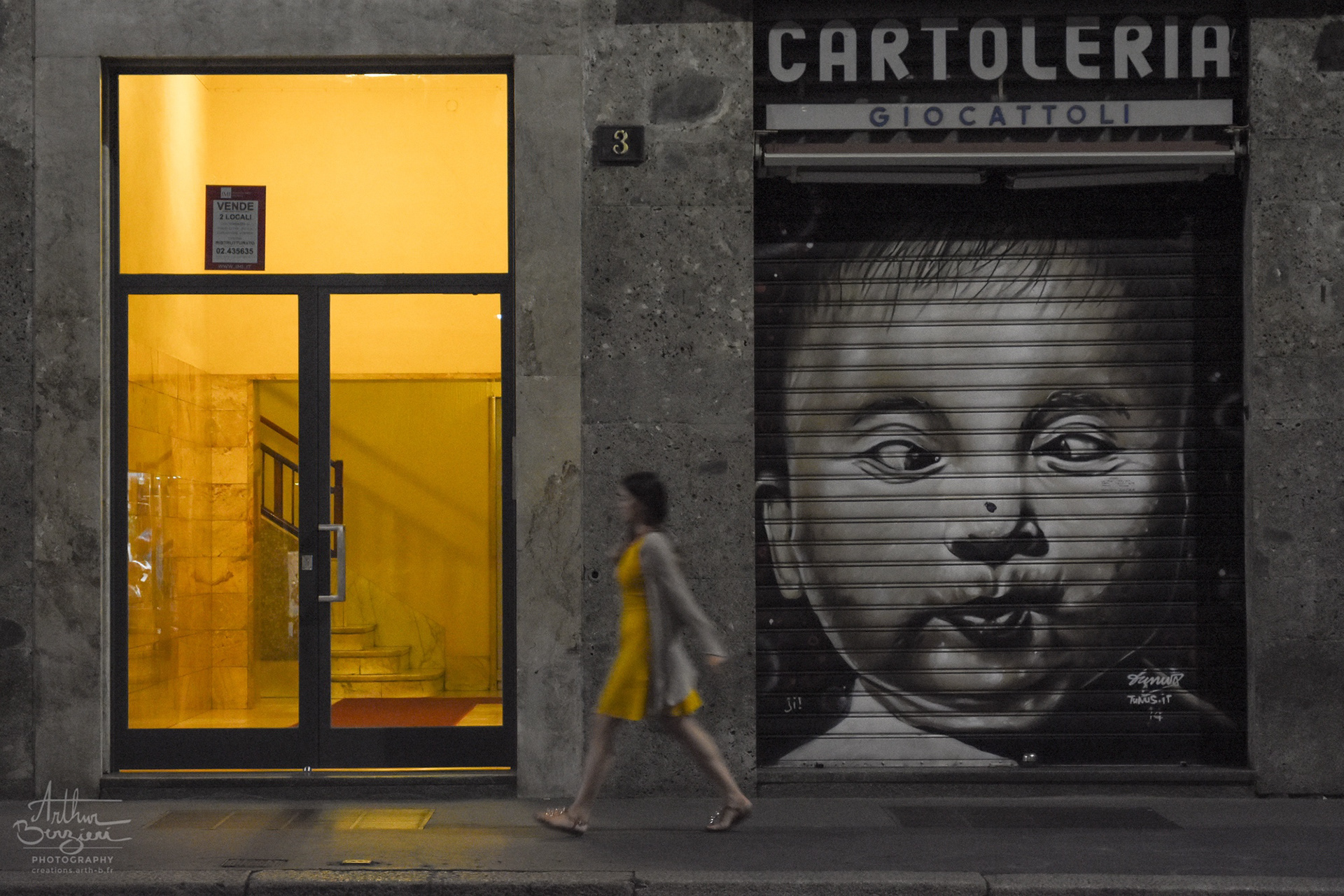 Cartoleria fermée et lumière chaude de l'entrée d'un immeuble qui semble colorer la robe de la jeune femme bien apprêtée.