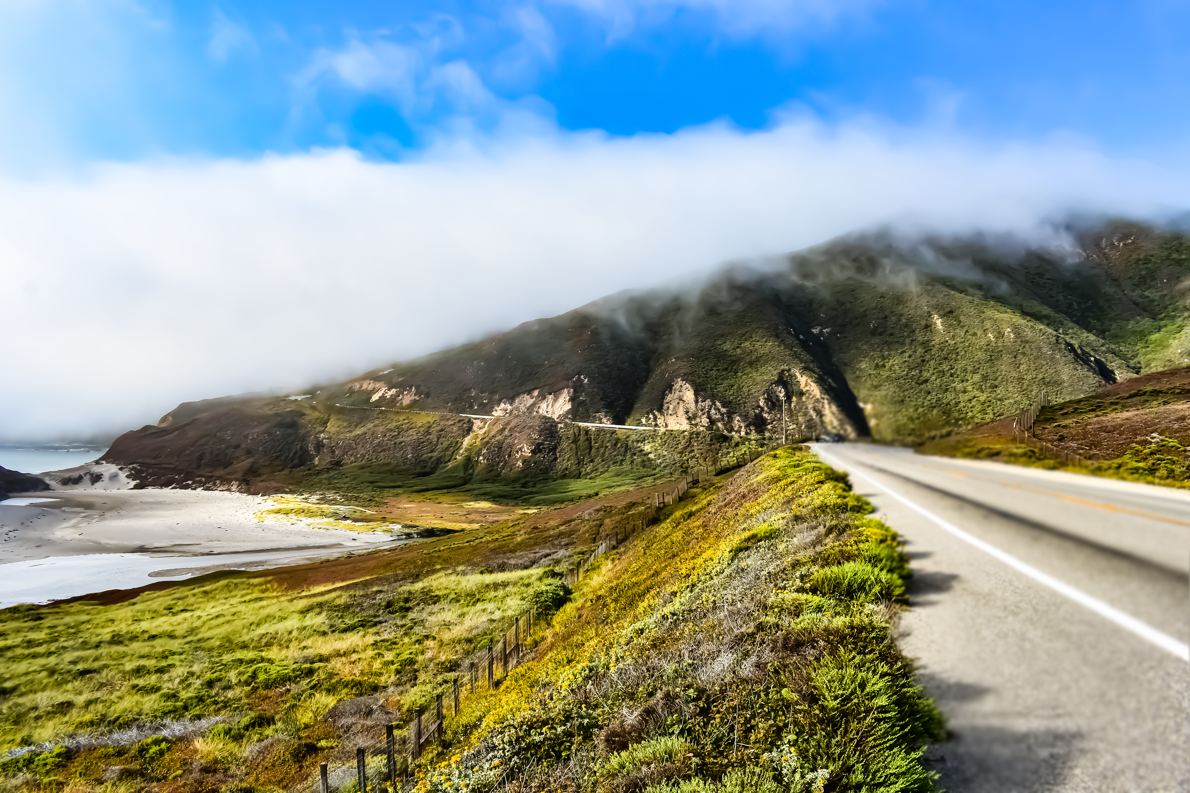 Panoramic landscape photograph showcasing the dramatic meeting of California's coastline, Highway 1, and rolling coastal cliffs.