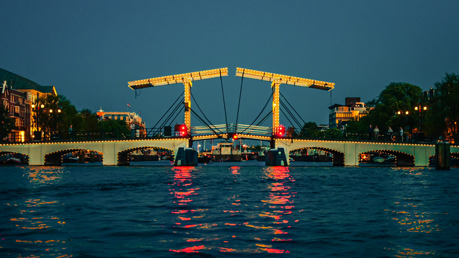 (ES) Este peculiar puente se encuentra en la pintoresca Amsterdam y cruza el río Amstel desde el año 1670. Según cuenta la historia, las hermanas Mager fueron las responsables de la construcción del primer puente que ocupó aquel lugar. Resulta ser que cada una de ellas vivía en uno de los lados del río, y para visitarse decidieron hacer frente a sus casas un puente. Pero era tan delgado que resultaba difícil que dos personas puedan transitarlo al mismo tiempo. De modo que, cuando el número de peatones que cruzaban el río mediante este puente se incrementó, el gobierno lo reemplazó por el actual, en 1871 aproximadamente.Es uno de los muchísimos puentes que fueron construidos en en esta ciudad, pero conserva ese algo especial que, sea por su mítica naturaleza o por su curiosa apariencia, no deja de hipnotizarte, sobretodo de noche cuando sus cientos de luces se reflejan en el rio. (EN) This quirky bridge is located in the beautiful Amsterdam and crosses the Amstel River since 1670. According to the story, the sisters Mager were responsible for building the first bridge that occupied that place. It turns out that each of them lived in one side of the river, and to be able to visit their homes, they built a bridge. But it was so narrow, that it was difficult for two people can walk at same time. Later, when the number of people crossing the river by this bridge was increased, the government has replaced it with the current one, in about 1871. It's one of the many bridges that were built in this city, but it retains something special that, by its mythical nature or their curious appearance, is impossible not to be mesmerized with its beauty especially when its hundreds lights reflect in the river at night.
