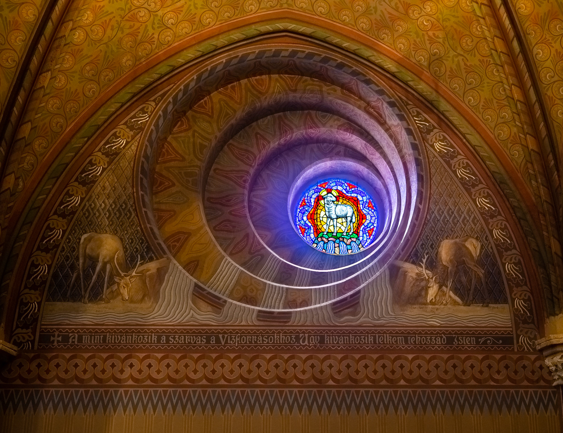 This circular window is above and to the right of a Neo-Roman baptismal fountain. The leftward skew of the spiral enables the circular opening on the inner surface of the wall to align with the baptismal fountain. The matched imagery of the four streams in the stained glass and through the spiral flow both visually and symbolically to the water-involving baptismal fountain. The lamb at the top of the hill from which the streams flow represents Jesus, the Lamb of God.