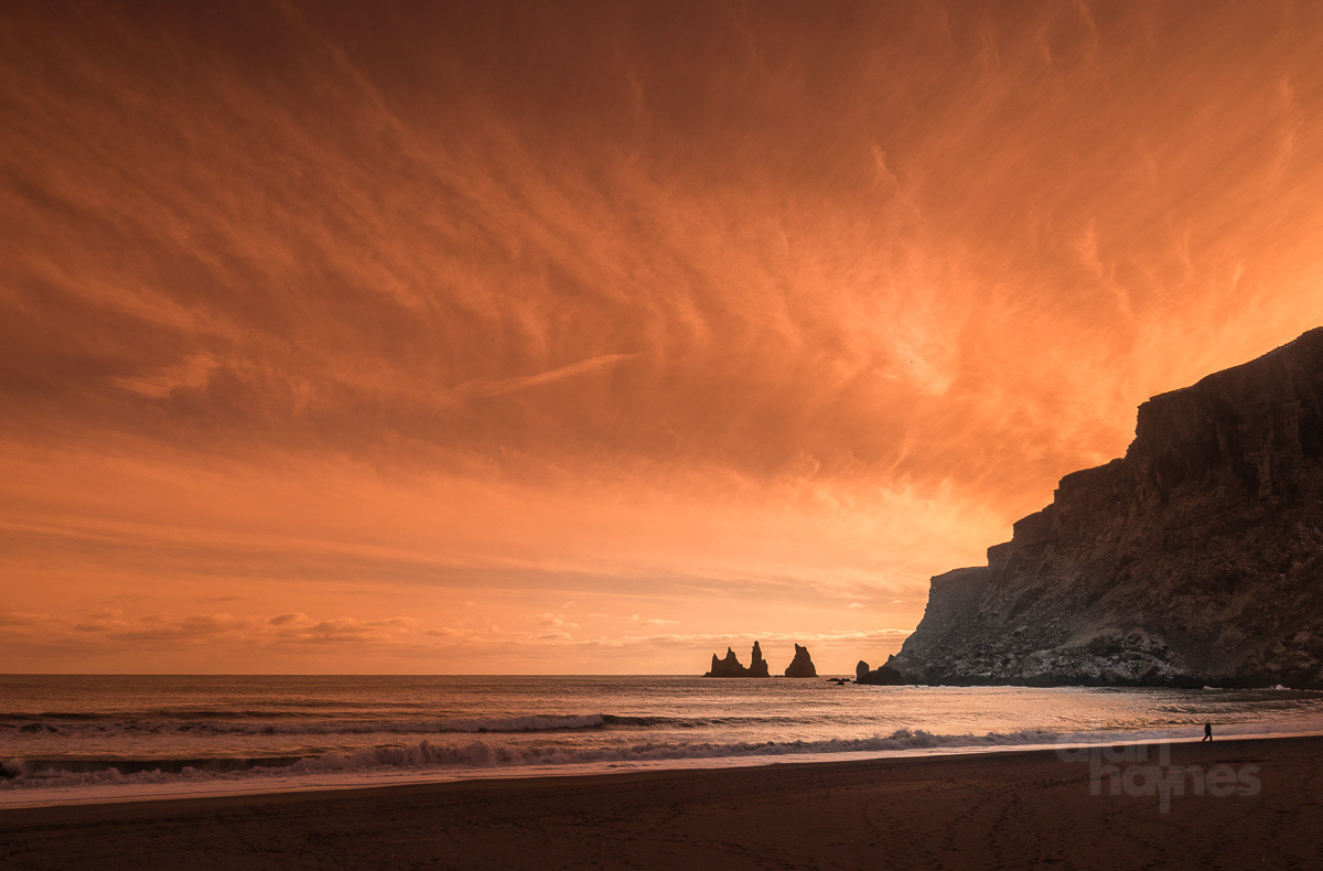 Man walking on the black sand beach at sunset with the Pinnacles of Vik in the distance. Vik, Iceland