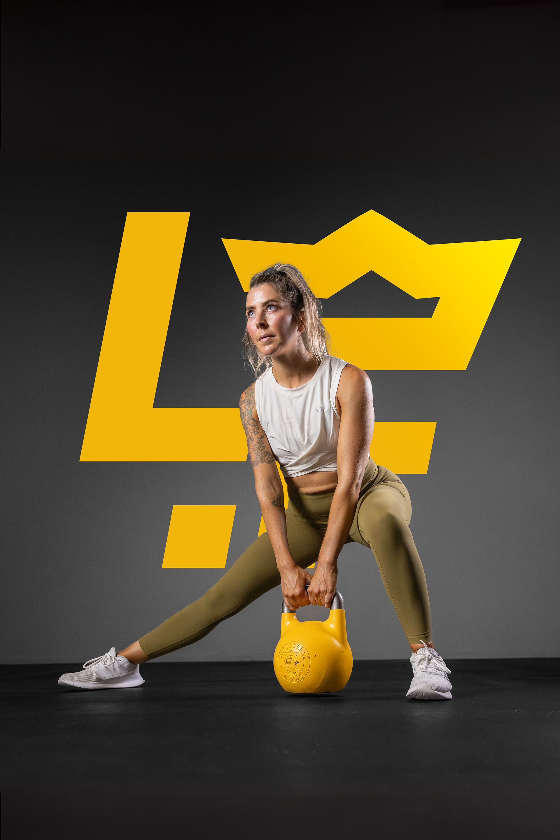 The image shows a woman performing a kettlebell exercise, standing in a wide stance while holding a yellow kettlebell with both hands. She is wearing a white sleeveless top and olive-green leggings, with a large stylized yellow crown logo in the background. The background is dark, highlighting the woman and the logo.