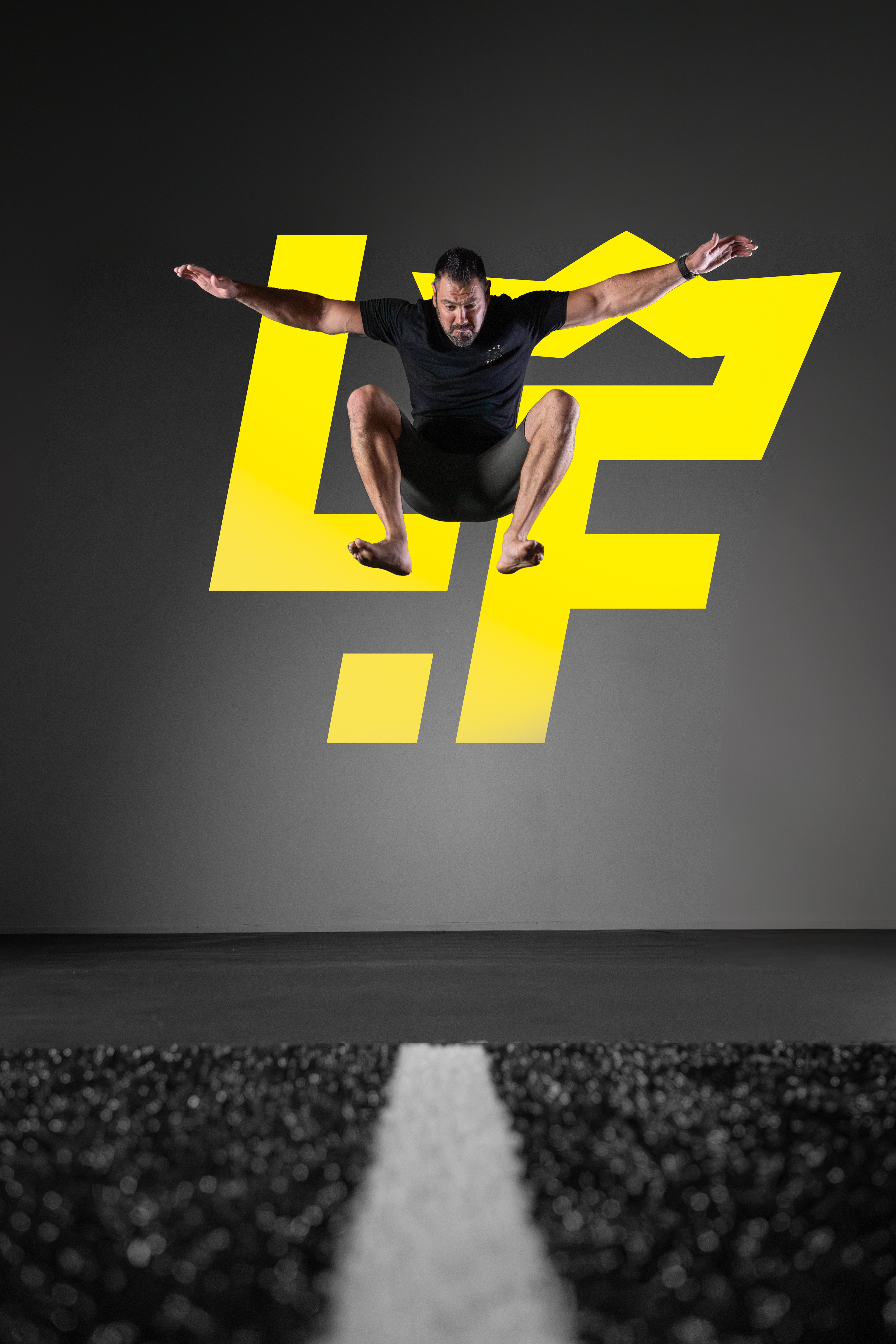 The image shows a man jumping high with his arms outstretched, wearing a black T-shirt and black shorts. He is positioned in front of a large yellow stylized crown logo against a dark background. The action and the athletic wear suggest a fitness or exercise-related theme.