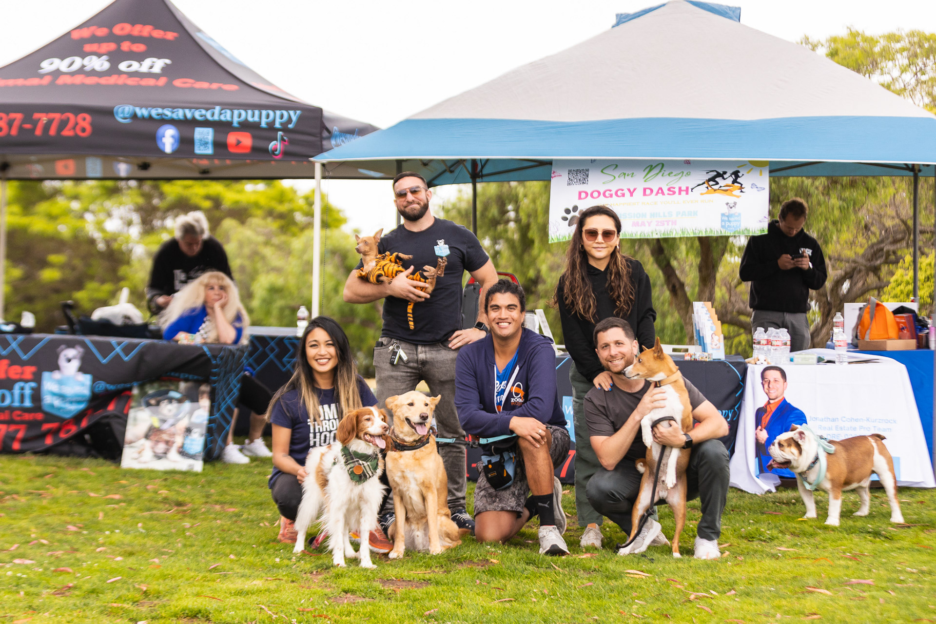 The image shows a group of people and their dogs posing at an outdoor event under a couple of tents. The backdrop includes banners with text such as "Doggy Dash" and "We Save Da Puppy." The people are smiling, and the scene suggests a community event focused on pets, featuring various dog breeds and promotional materials for pet care services.