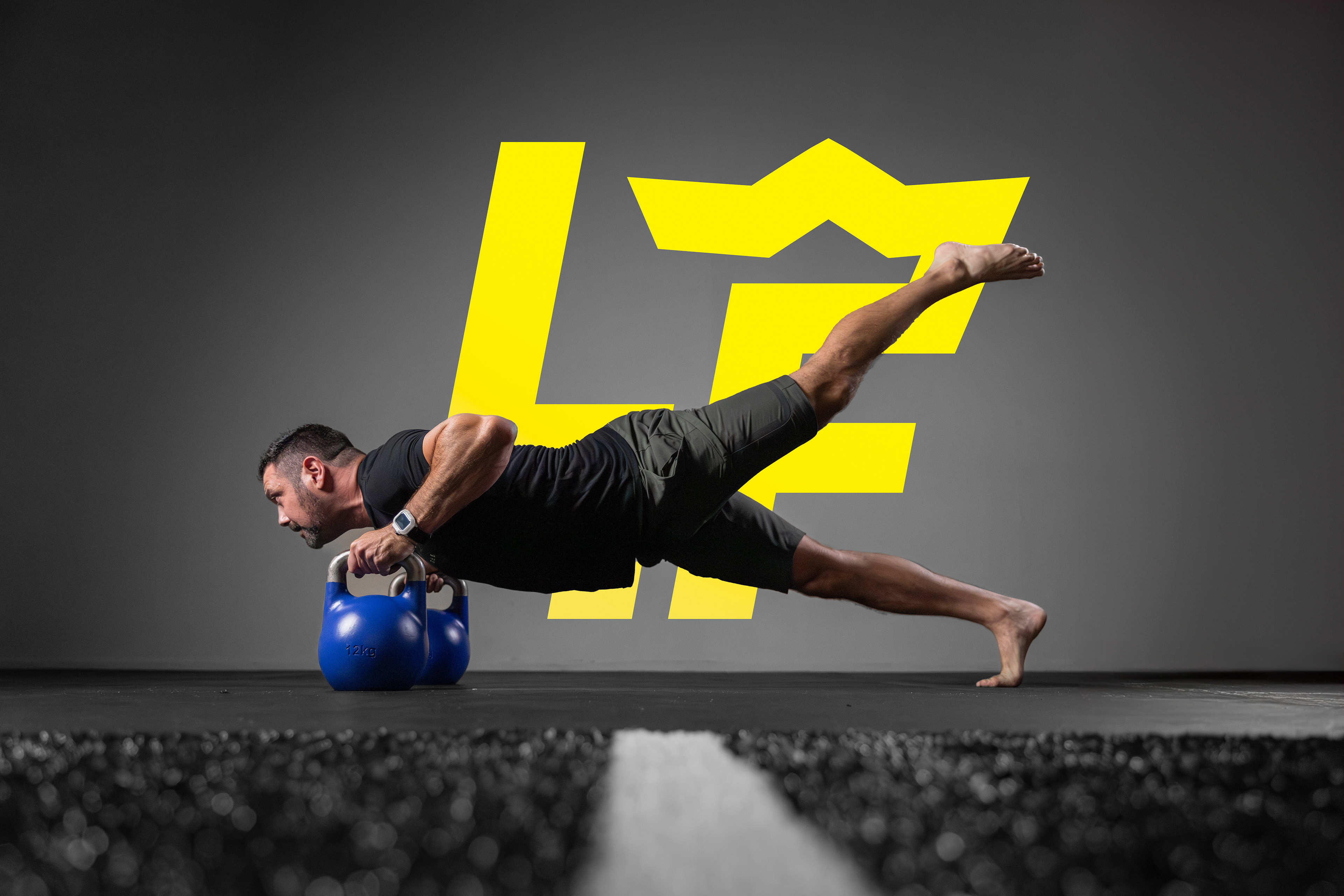 The image depicts a man performing a challenging kettlebell exercise, holding two blue kettlebells while balancing on one foot in a plank position. He is dressed in a black T-shirt and shorts, with a large yellow stylized crown logo in the background. The dark setting and intense posture highlight a focus on fitness and strength training.