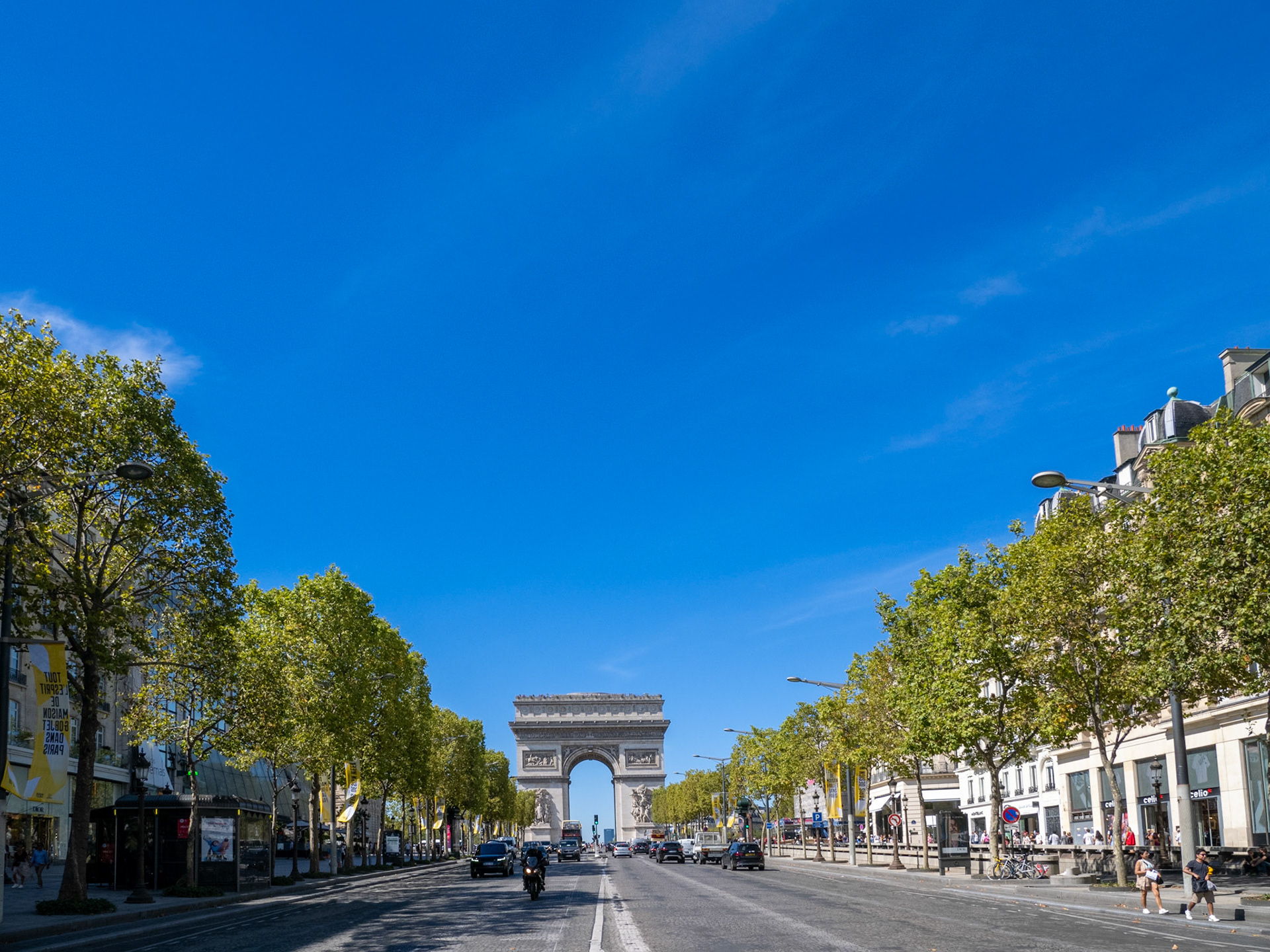 From street level, pointing up into a large, blue summer sky is a perspective view of the Arc de Triomphe and Champs-Élysées