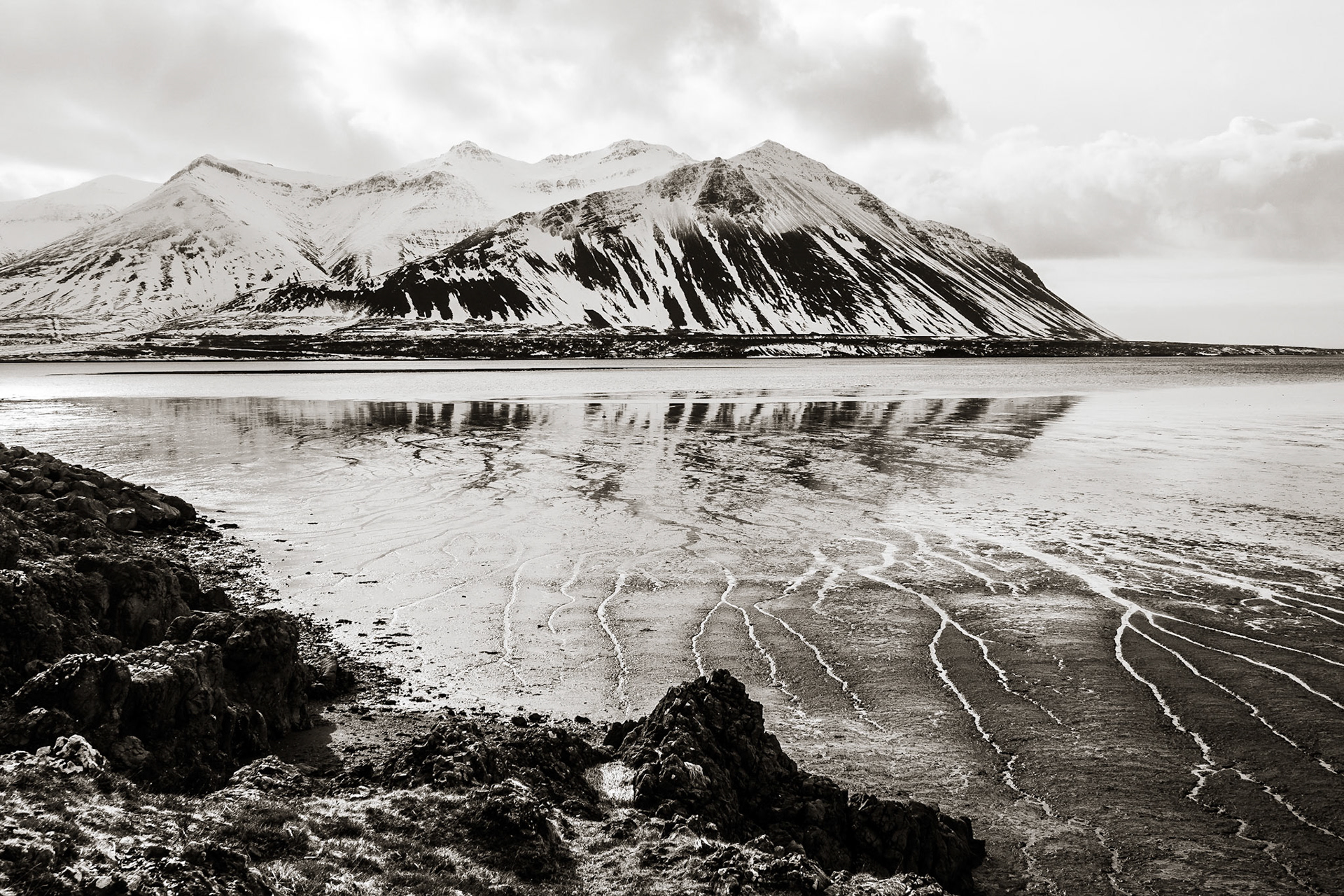 Dramatic snow-covered Hafnar mountain rises out of the Borgarnes fjord at low tide with rivulets etched into the mud of the seafloor along a rocky coastline.