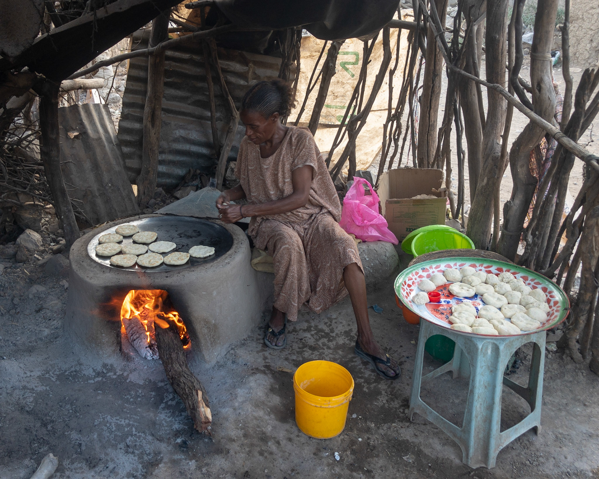 An Ethiopian woman makes and bakes small flat-breads over a flat iron-topped outdoor wood stove in a ramshackle enclosure of sticks, branches and corrugated iron sheeting.