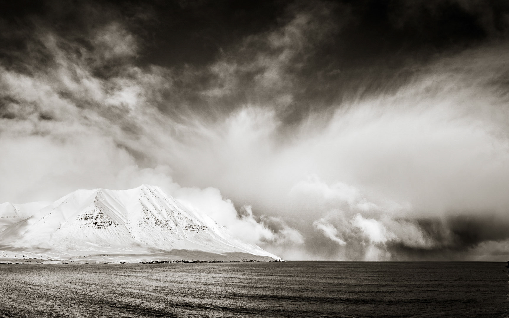 Dramatic storm clouds build off the coast of a fjord in Iceland with bright white snow-covered mountains plunging into the dark sea under a blue sky with wispy clouds.