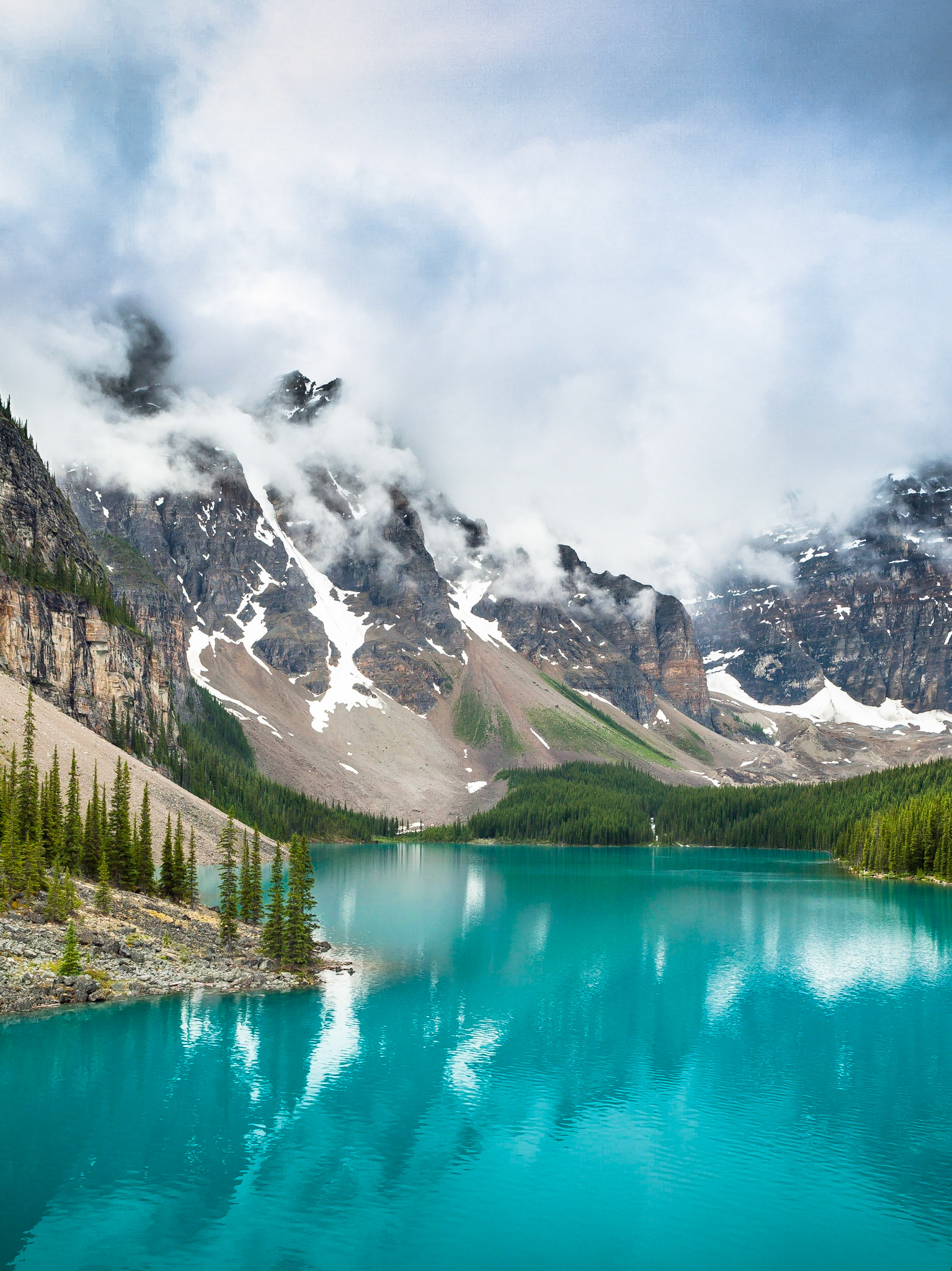 The spectaular tourquoise blue glacial waters of Moraine Lake shimmer against the dramatic cloudy backdrop of the Rocky Mountains in Banff National Park, Alberta, Canada