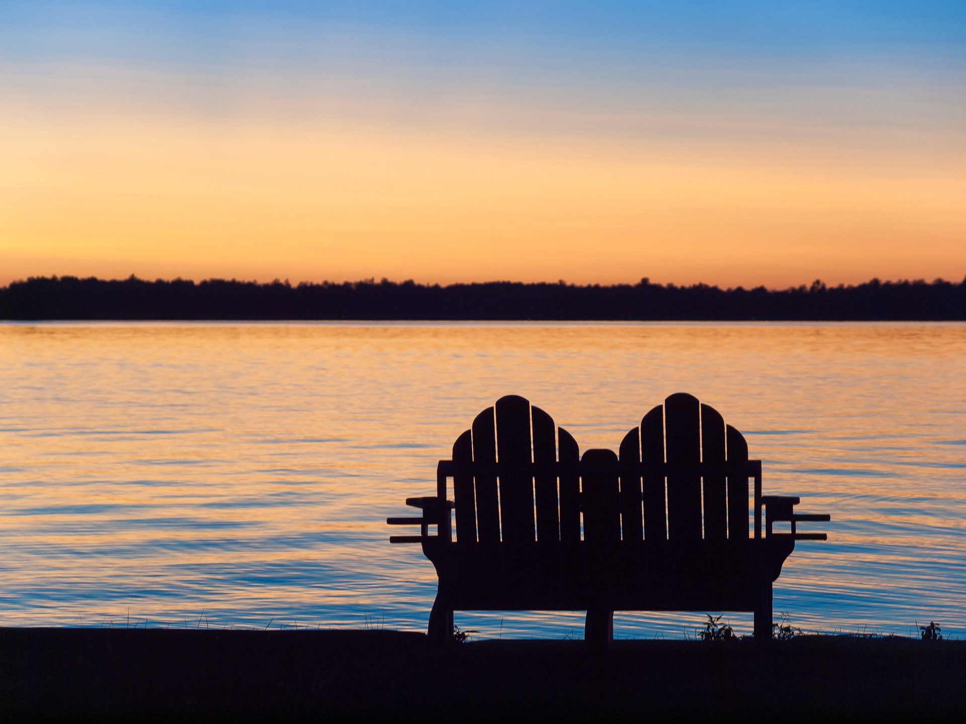 A double wooden adirondack chair for two is silhouetted against the colours of sunset along the shore of a lake.