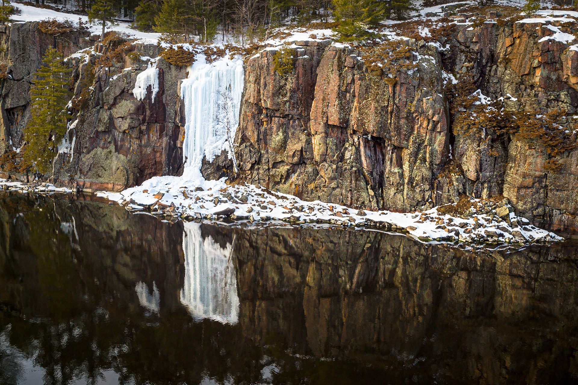 A white frozen waterfall spills down a dark pink-brown sheer cliff of rock reflected in a dark placid river