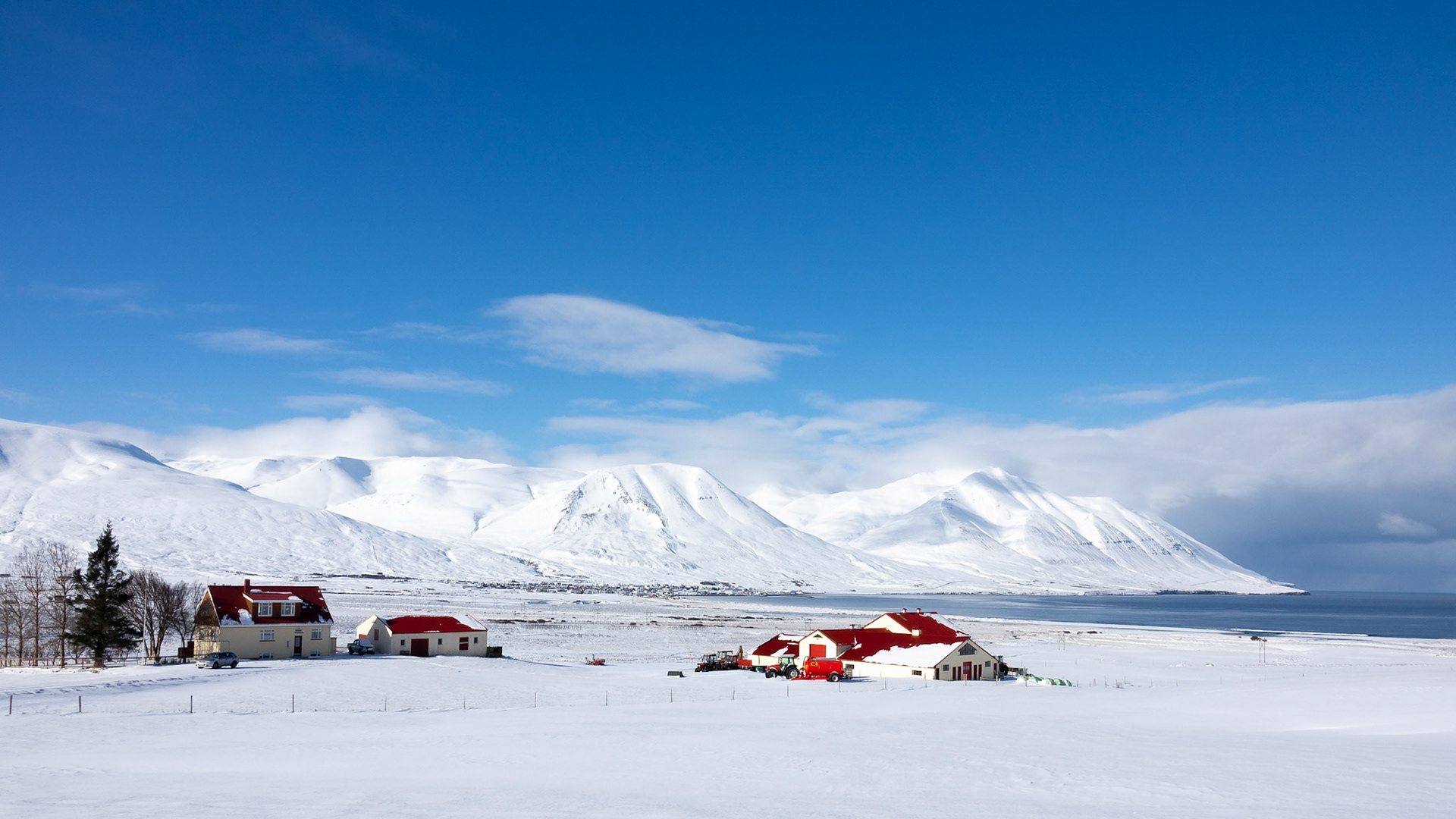 A pale yellow farm house and outbuildings with red roofs sits amongst the snow-covered fields along a dramatic fjord coast of mountains and blue ocean.