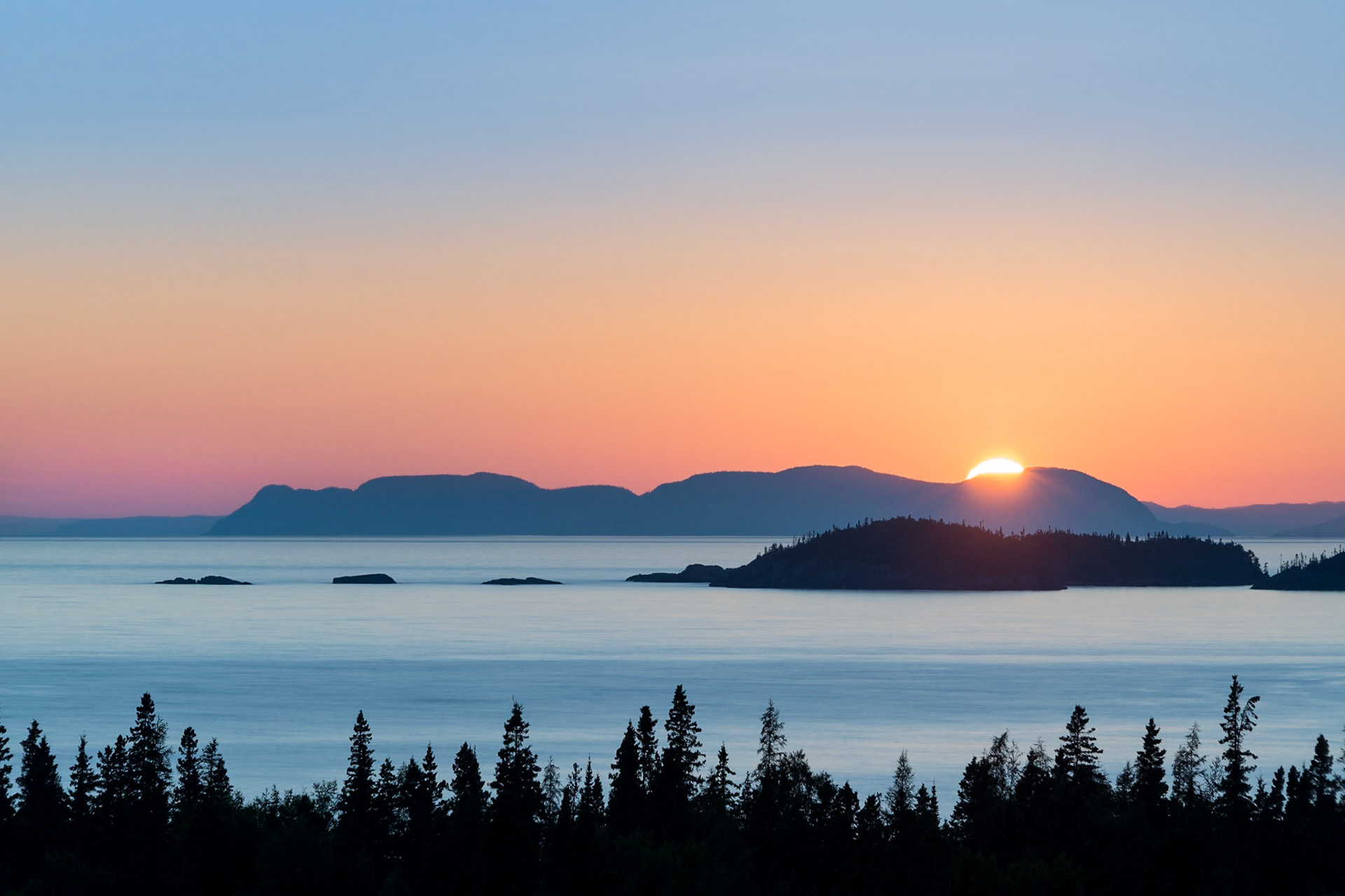 The setting sun kisses the undulating and irregular profile and rocky promontory of Pic Island along the north coast of Lake Superior with islands and the silhouette of the boreal forest in the foreground.