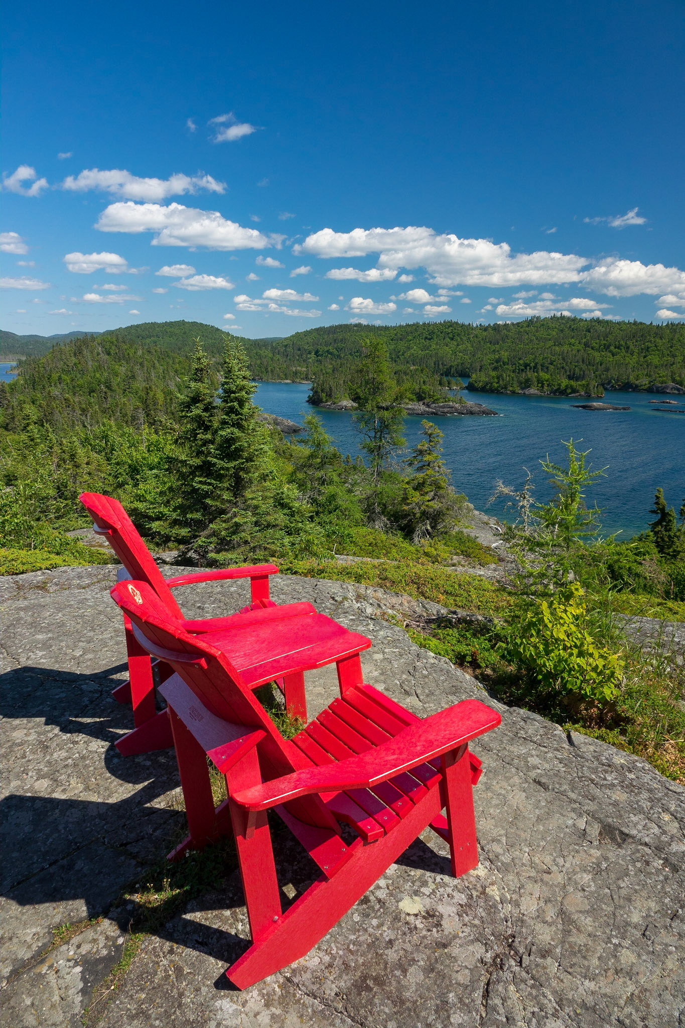 Two right red Parks Canada Adirondack chairs are on a rocky outcrop high above the deep blue waters of Lake Superior with distant views of the rich green boreal forest all around.