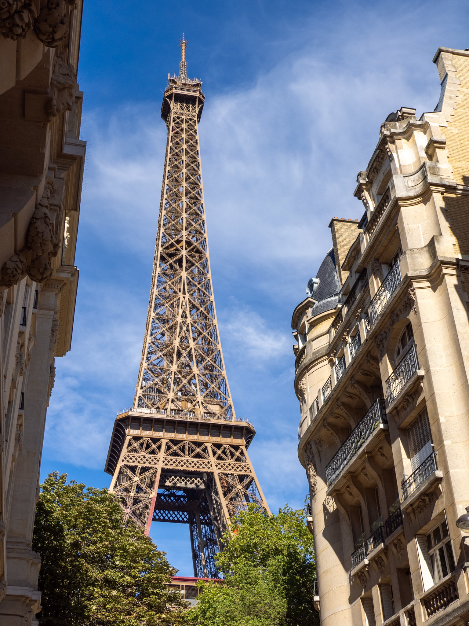 The Eiffel Tower looms above green trees and a block of apartments in thei narrow street view looking up into a blue sky with clouds.