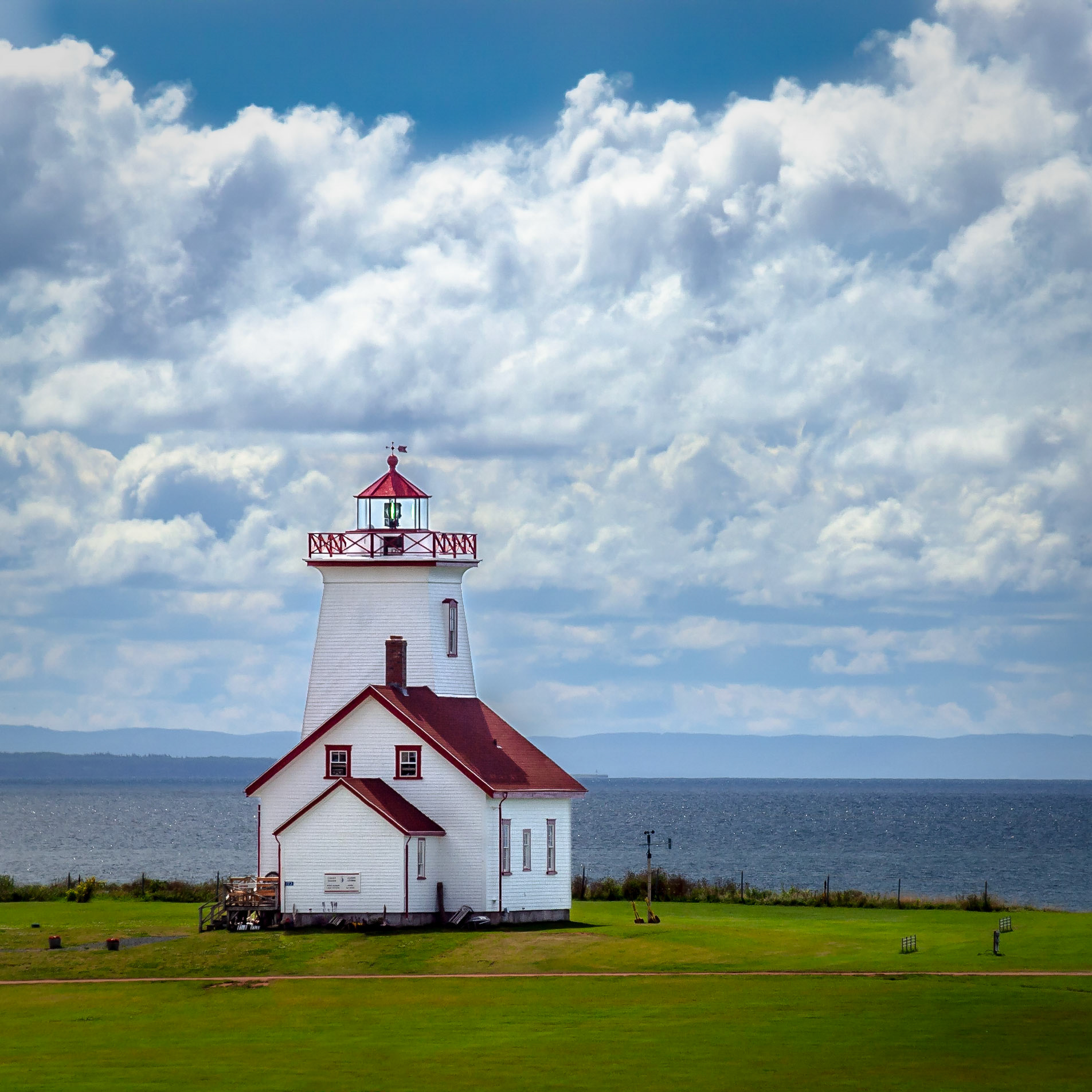 Brightly backlit white and red lighthouse surrounded by green grass looking out over the sea with Nova Scotia on the far shore and billowy clouds in the blue sky