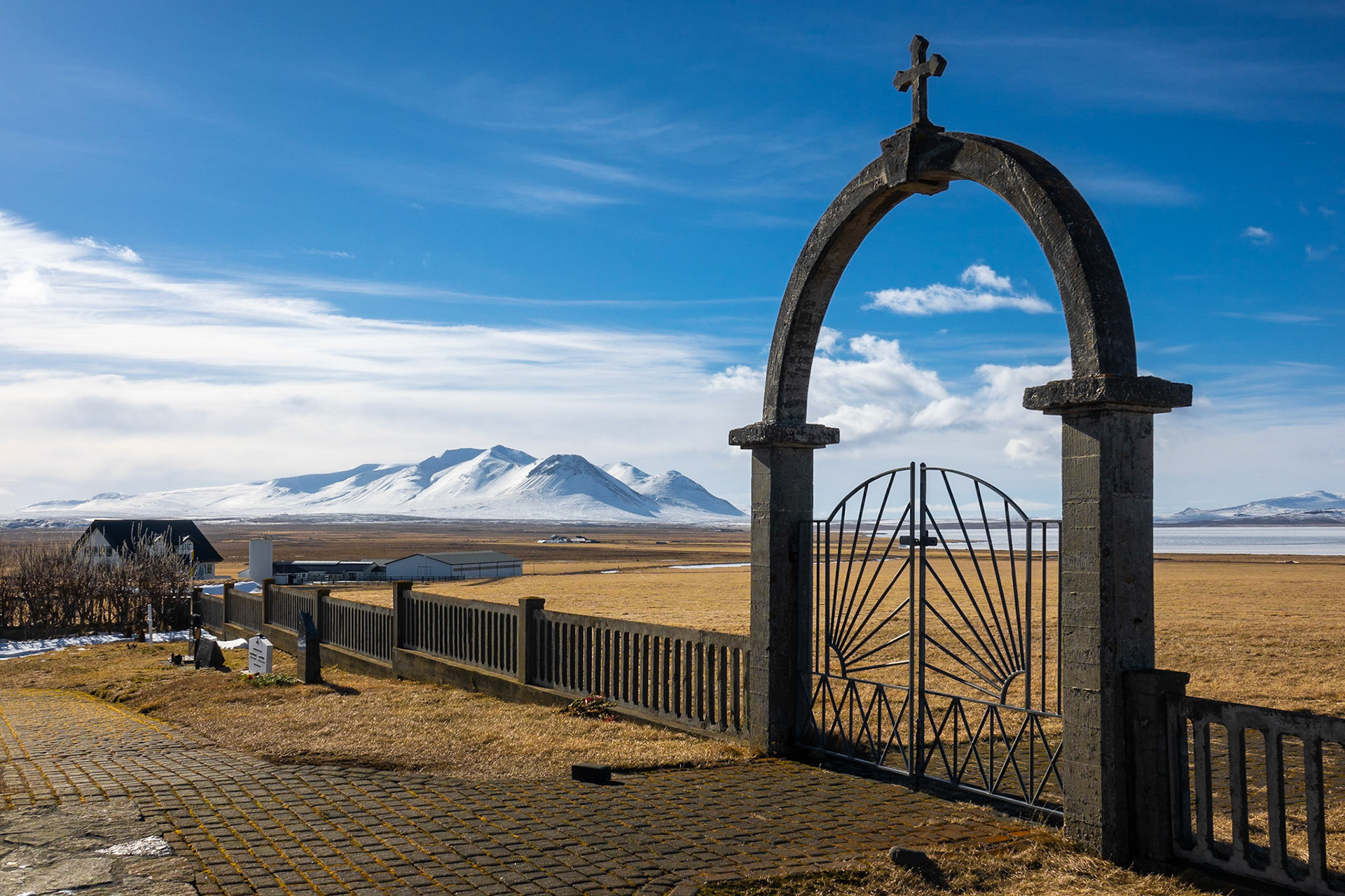The black basalt arch, gate and stone fence of the entryway into the church yard sits under a brilliant blue sky with snow-covered mountains in the background with wind-swept fields between.