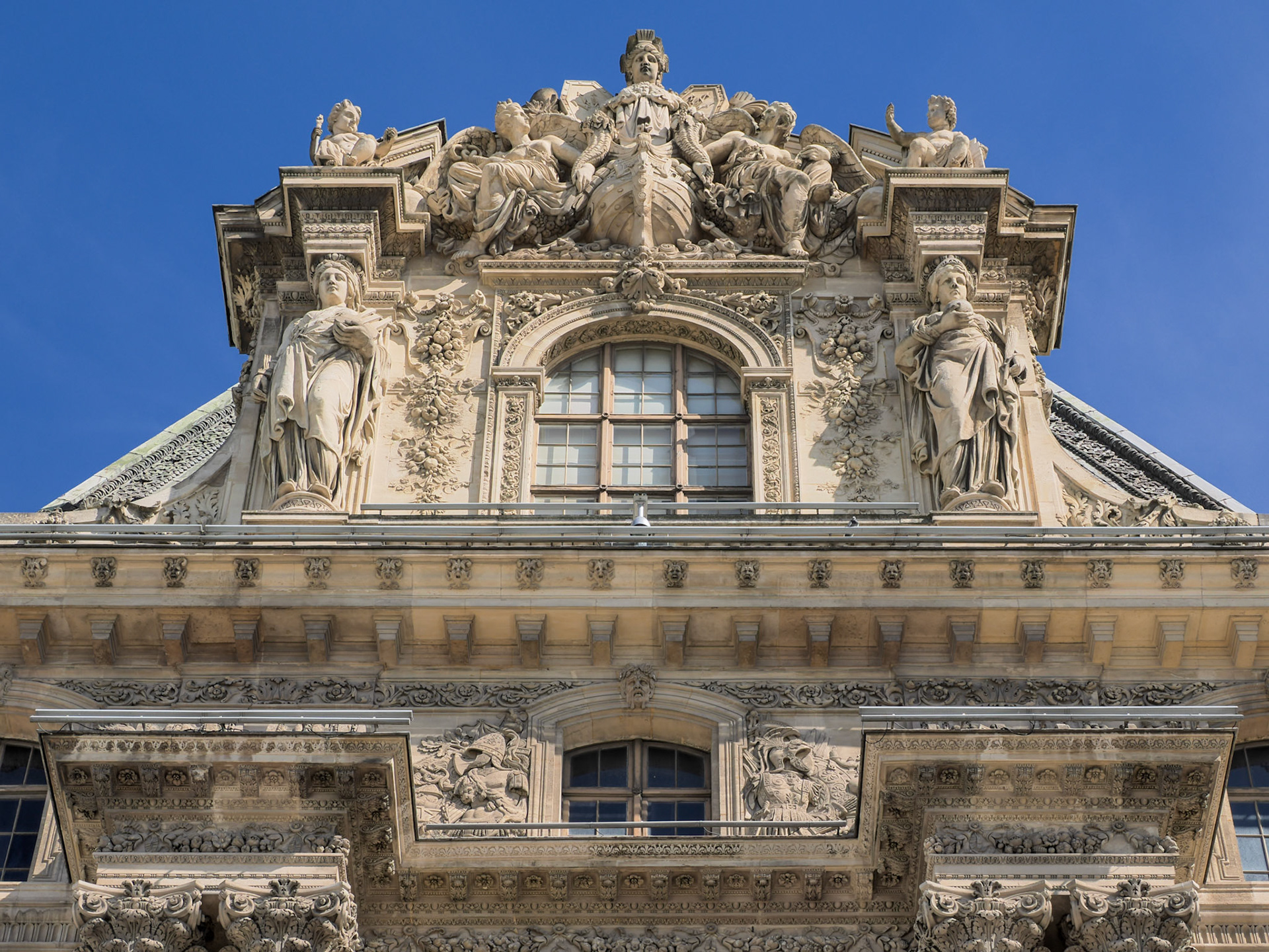 An oblique view from the ground of exquisitely detailed ornate carvings in stone adorn the front of part of the Louvre Museum, Paris, with a clear blue sky behind