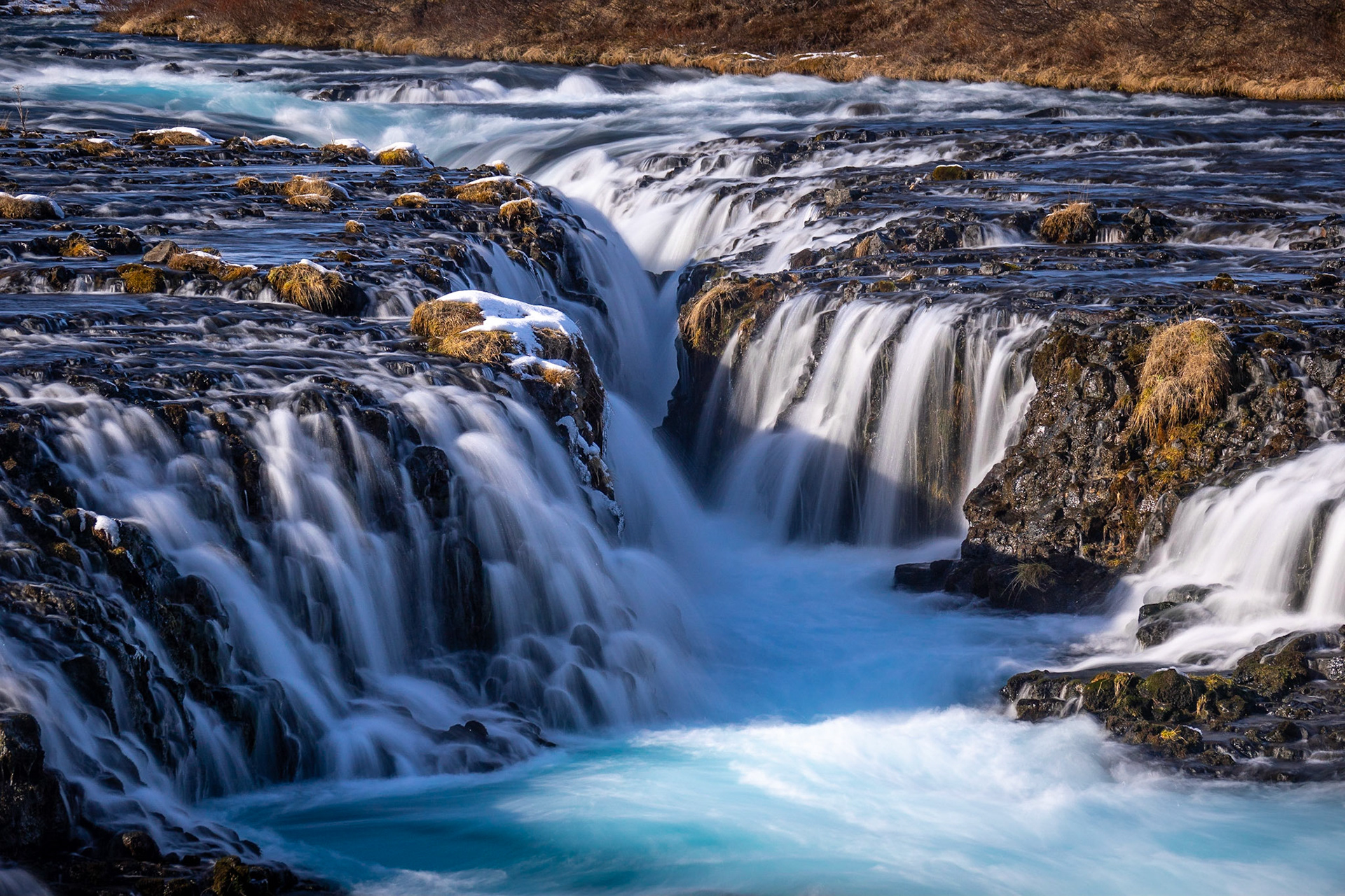 A detailed photo of the the many cascades and rivulets of waterfalls with powder blue and turquoise glacial water flowing over black lava rock and through a steep-sided fissure in the river bed.