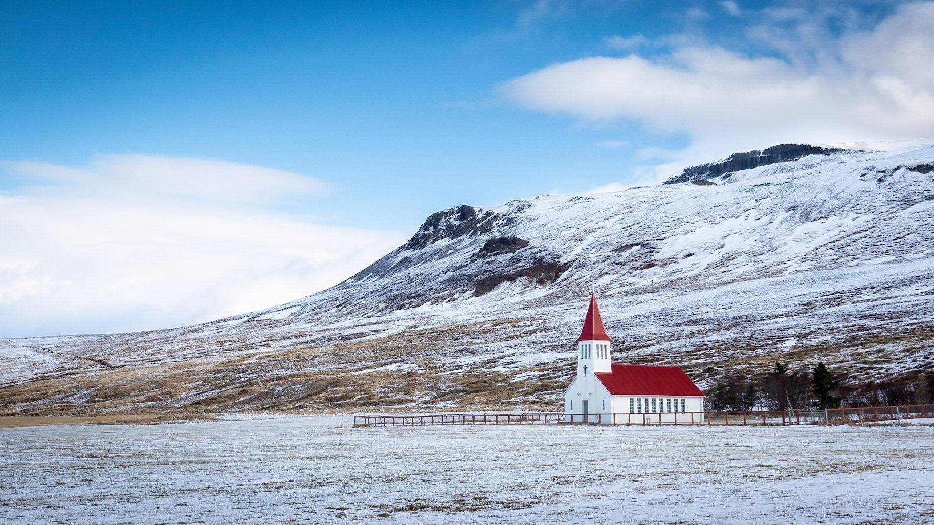 The white building with red roof church of Hoskuld stands out in a wide field against a back drop of snowy mountains under a blue sky with scattered clouds.