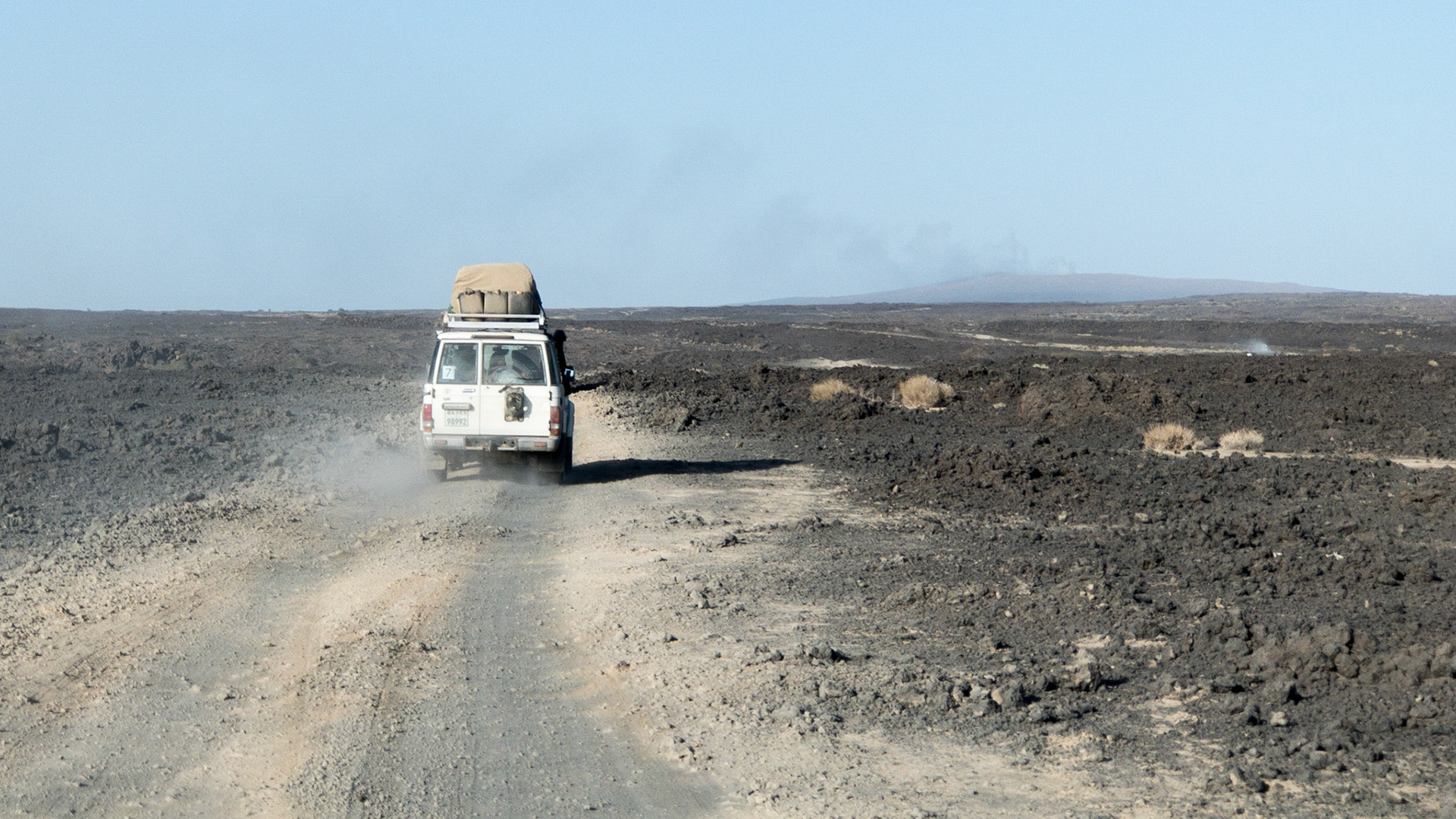 An expedition truck drives across a very rough, grey lava field with Erte Ale volcano in the background, in Ethiopia's Danakil Depression, the hottest, driest, most inhospiotable place on Earth.