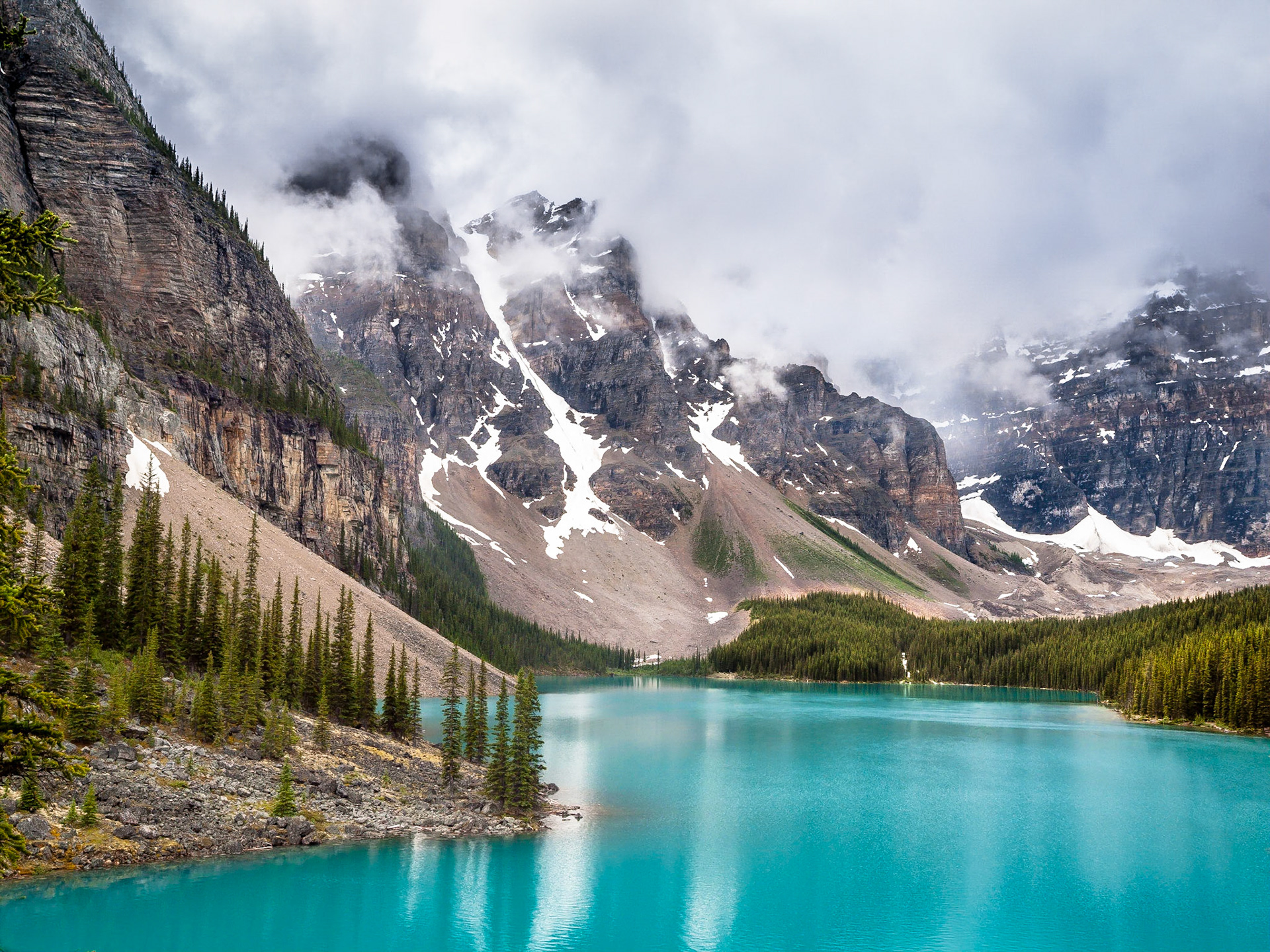 The spectaular tourquoise blue glacial waters of Moraine Lake shimmer against the cloudy backdrop of the Rocky Mountains in Banff National Park, Alberta, Canada