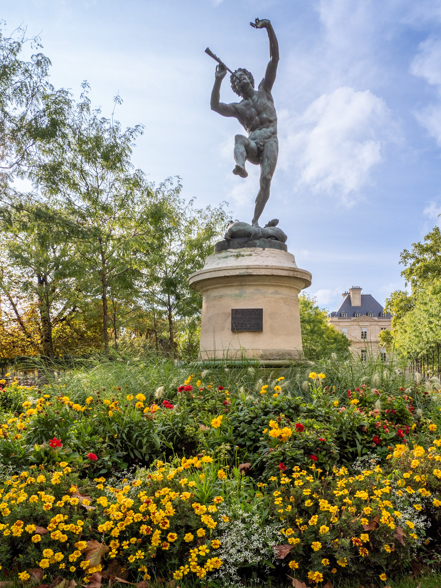 A bronze statue of Faune (or Faunus) enthusiastically playing his flute is surrounded by late summer flowers and trees against a pale blue sky with some light cloud. Statue by Eugene Louis Lequesnes.