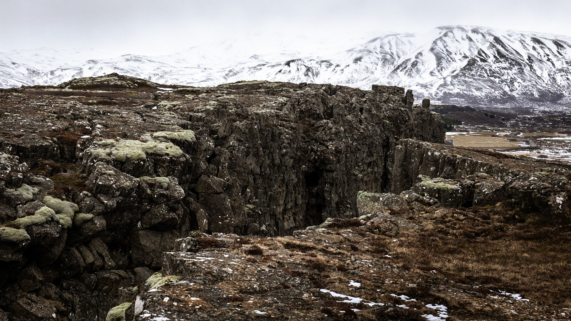 A wide fissure cuts diagonally through the dark, black basalt lava field of Thingvellir -
 a UNESCO World heritage Site - with snow-covered mountains in the background.