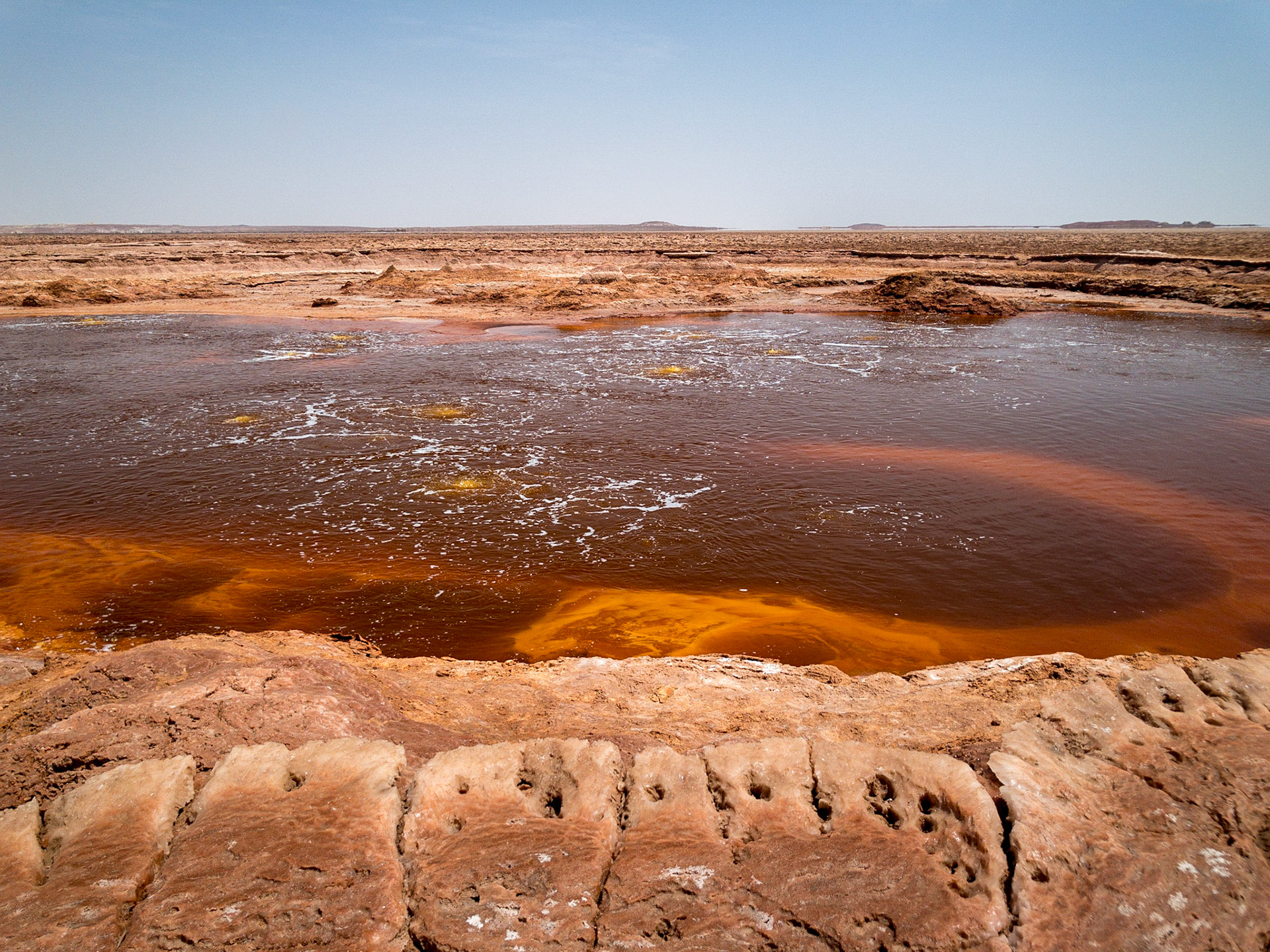 The clear, brown waters of a sulphuric acid lake bubble and churn creating orange-brown banks of salt on a flat landscape in the Danakil Depression, Ethiopia, the hottest, driest, most inhospitable place on Earth.