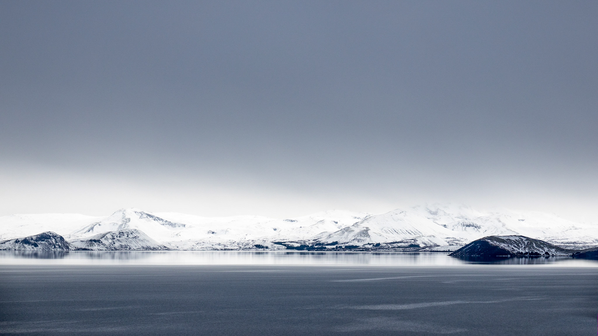 A sombre, steel-grey cloudy sky hovers over Thingvalla Lake along the rift valley of Iceland with snow-covered mountains along the shore