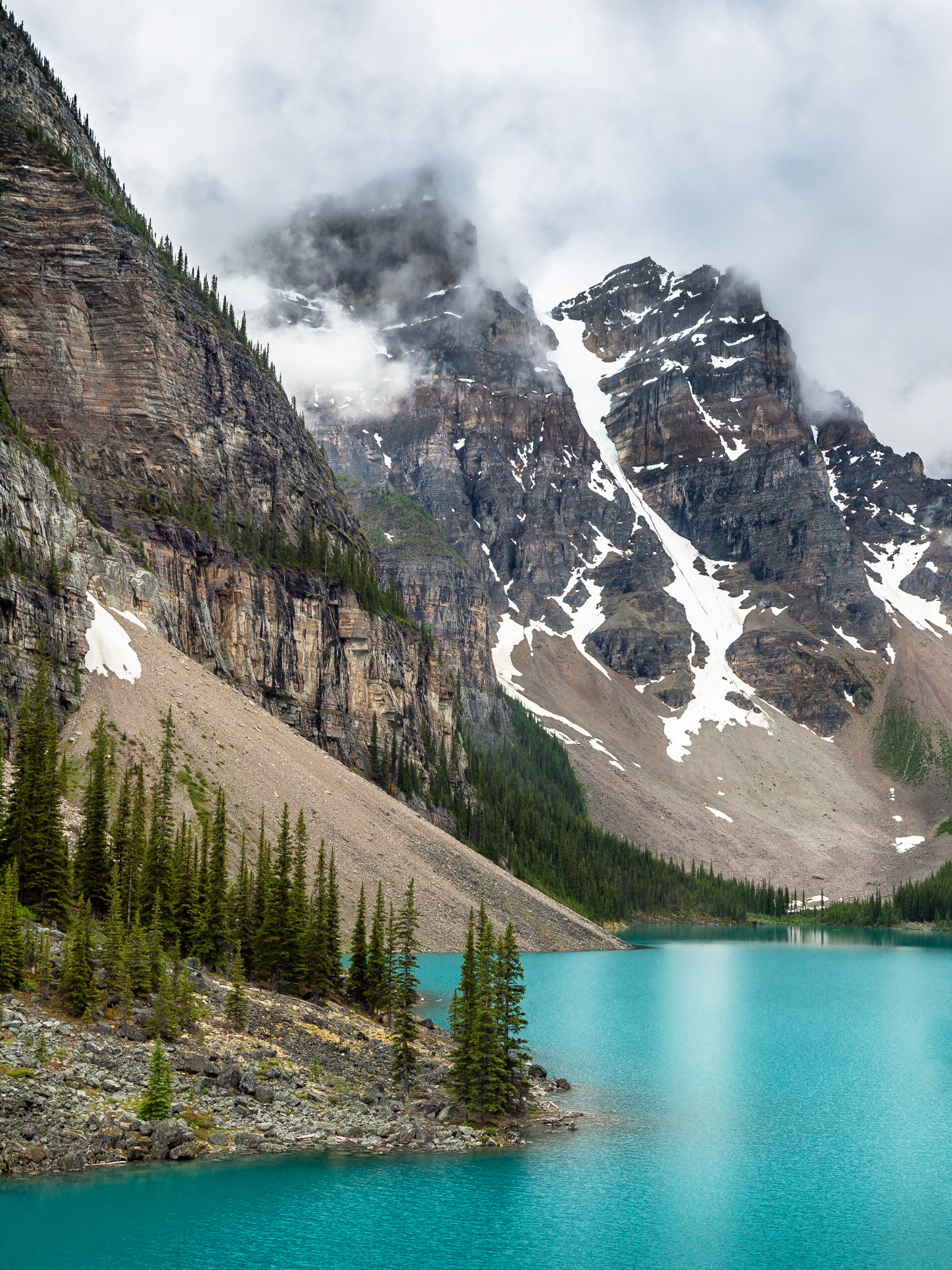 The spectaular tourquoise blue glacial waters of Moraine Lake shimmer against the cloudy backdrop of the Rocky Mountains in Banff National Park, Alberta, Canada