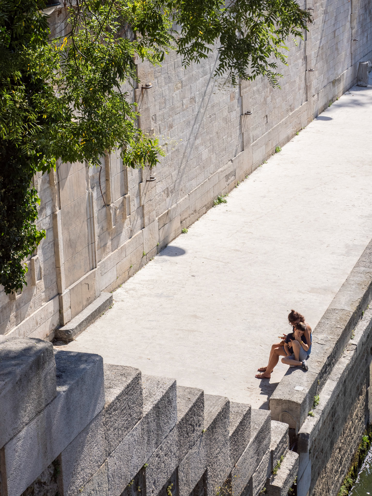 A small boy and his mother sit on a stone wall on the stark, white stone quay along the River Seine, Paris