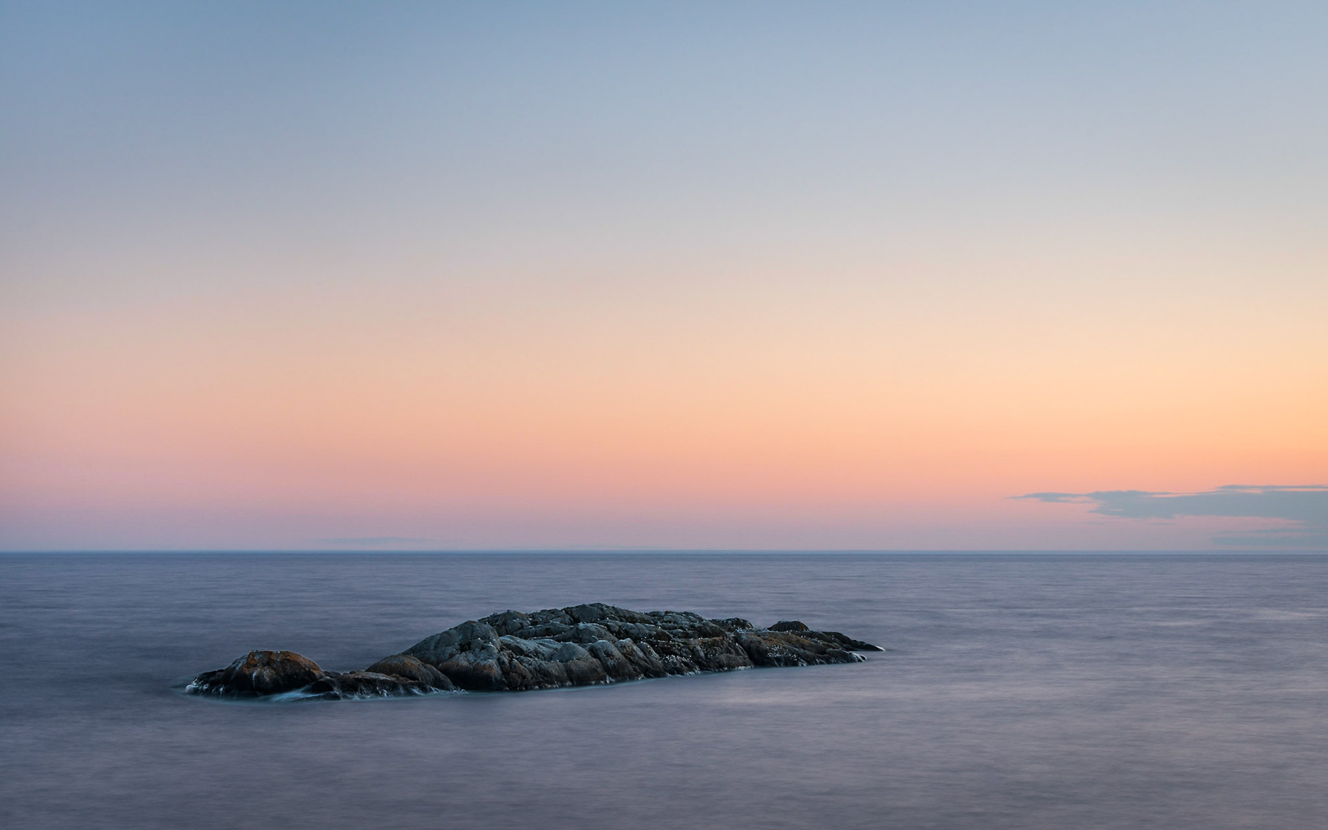 The pink and magenta light of dusk warms the sky above a cold, lone rocky outcrop surrounded by the broad, blue expanse of Lake Superior.