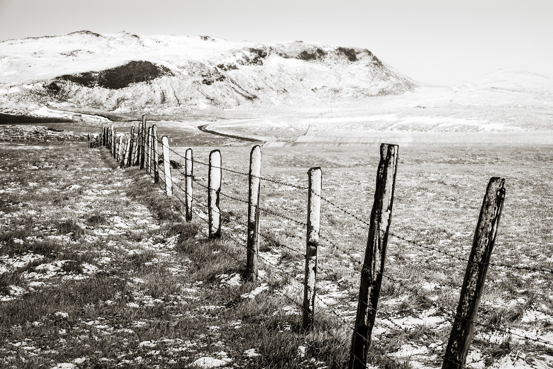 Hand-hewn pickets of a fence line are etched in snow as they march across an abandonned field with snow-covered mountains behind